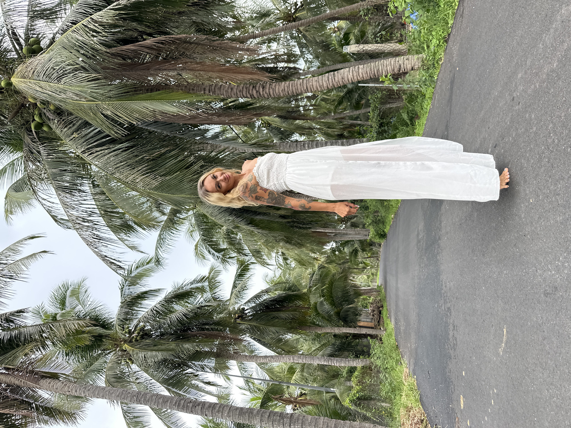 In Mũi Né, Vietnam, a woman with blonde hair and tattoos on her arm stands on an asphalt road lined with numerous tall palm trees. She is wearing a white, strapless maxi dress. The trees are abundant, with coconuts visible on some, creating a tropical atmosphere. The road appears to be a pathway or street surrounded by lush vegetation and is empty of other people or vehicles. The sky is overcast.