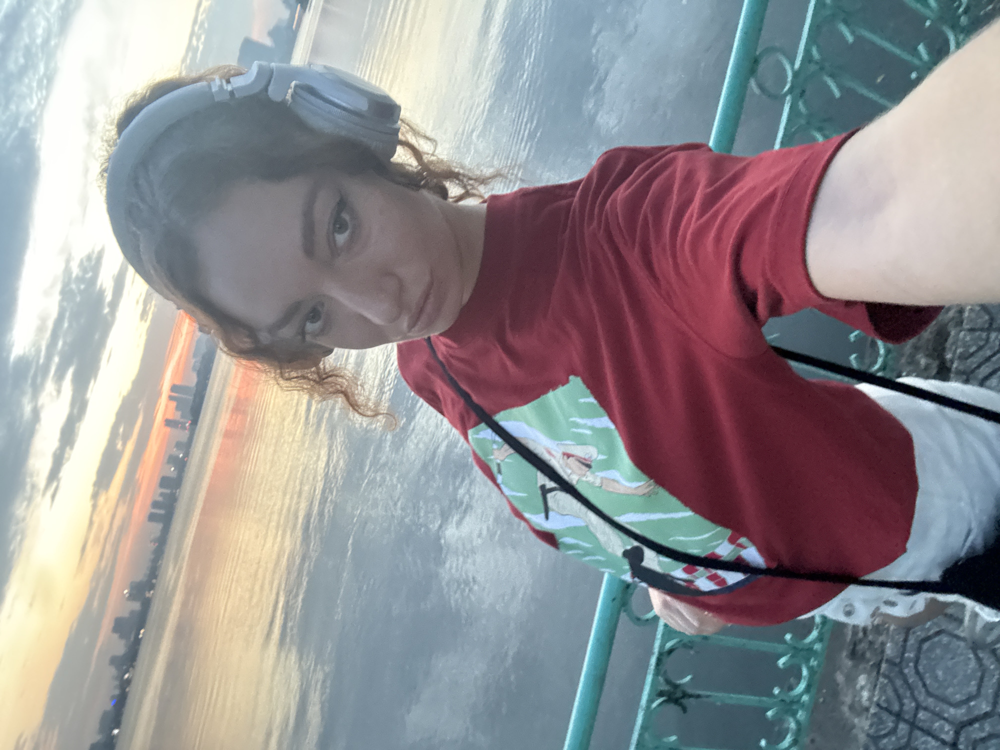A young woman with curly brown hair and headphones is taking a selfie on a walkway next to a body of water. She is wearing a red t-shirt with a graphic print on the front and white shorts. The water reflects the sky, which is filled with clouds and hints of an orange and yellow sunset. In the background, a city skyline is visible. The walkway has a teal-colored railing. The scene appears to be located on West Lake in Hanoi, Vietnam.