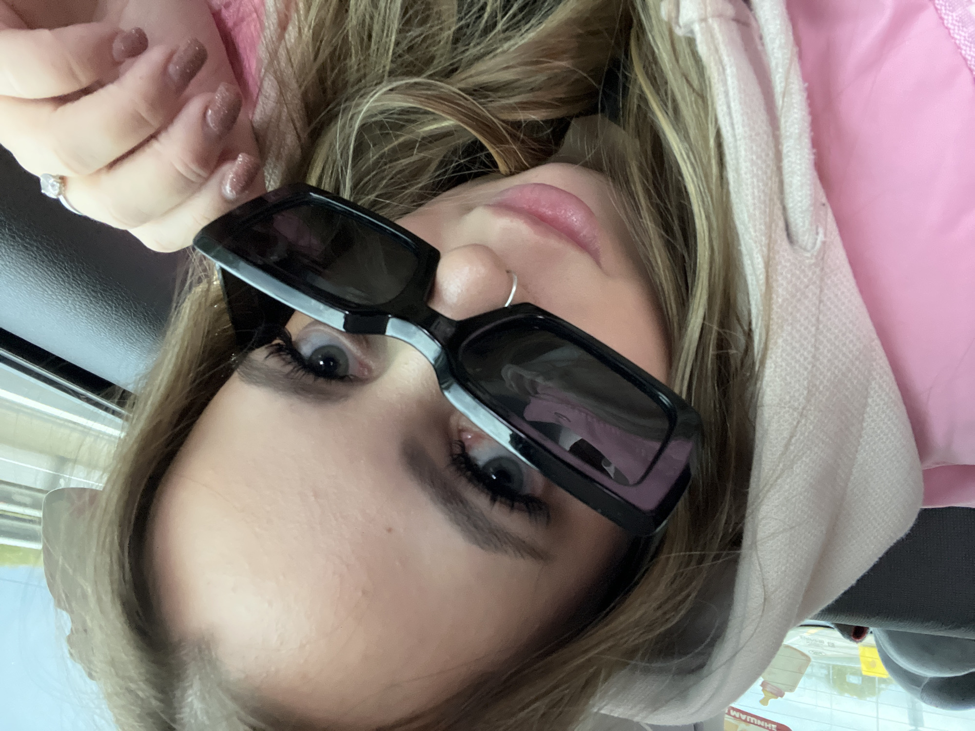 A woman with fair skin, blonde hair, and blue eyes is wearing black rectangular sunglasses and a pink hoodie. She has a silver nose ring and a diamond ring on her left ring finger. Her fingernails are painted with a glittery taupe polish. A partial view of an exterior scene with a window or windshield reflection, including a sign with Cyrillic letters indicating a possible location in Saratovskaya, Russia, is visible.