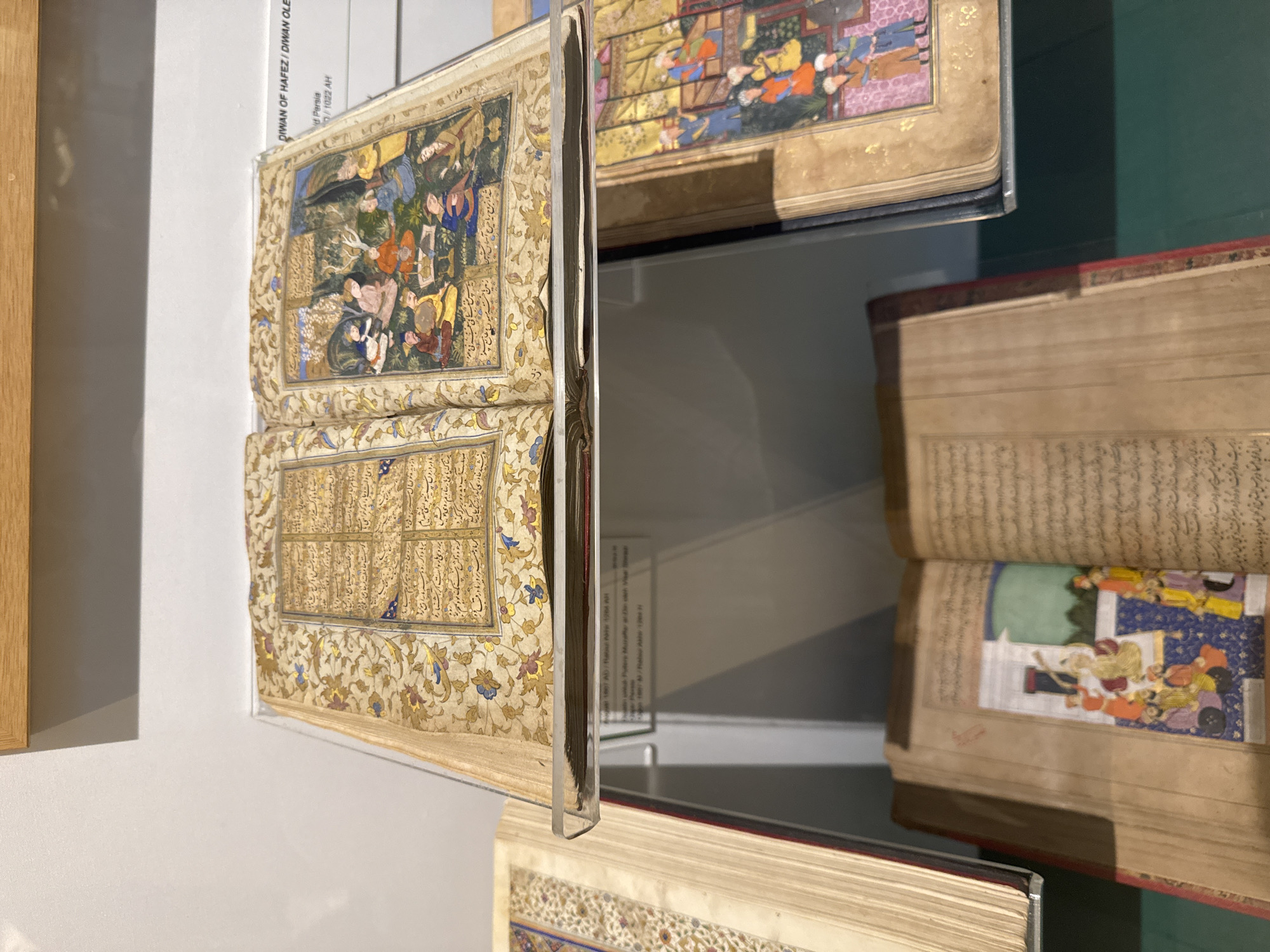 The image shows an open book displayed in a museum or gallery setting, likely in Kuala Lumpur, Malaysia. The book, identified by the label "DIWAN OF HAFEZ/DIWAN OLE," appears to be a historical or cultural artifact. The open pages exhibit intricate artwork and calligraphy, with detailed miniature paintings of people and scenes. The pages also contain text in a script that is likely Arabic. The book is displayed in a transparent protective case, and other similar books are visible on shelves or displays behind it. The overall environment appears well-lit and professional, suggesting a controlled museum or gallery setting.