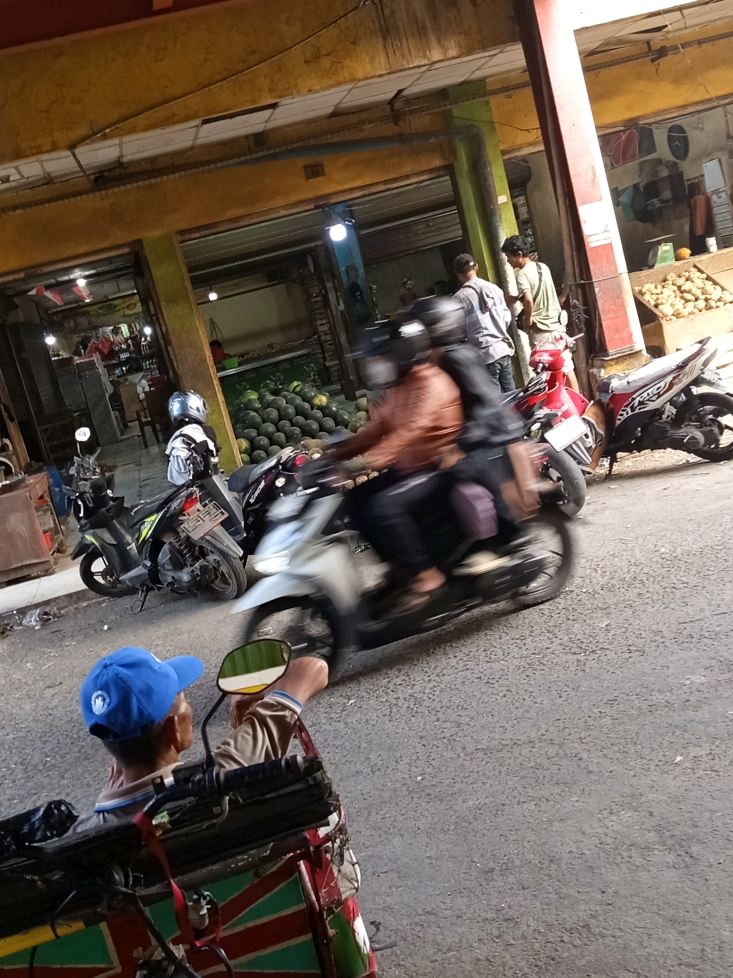In Karawang, Indonesia, a bustling market scene unfolds under an overhanging structure with a yellow roof and beams. In the foreground, a man wearing a blue baseball cap is seated in a motorized tricycle, facing away from the camera and towards the market. He is looking at a motorcycle that is blurring past, with a rider wearing a brown jacket and a helmet. Behind this motorcycle, another motorcycle, a red one, is parked. Further into the market, several other motorcycles are visible, including a black one in the middle ground. Stalls are set up within the market, displaying produce such as watermelons and potatoes. People, some in casual attire, are seen walking and interacting within the market environment. The overall impression is one of a busy and active marketplace.