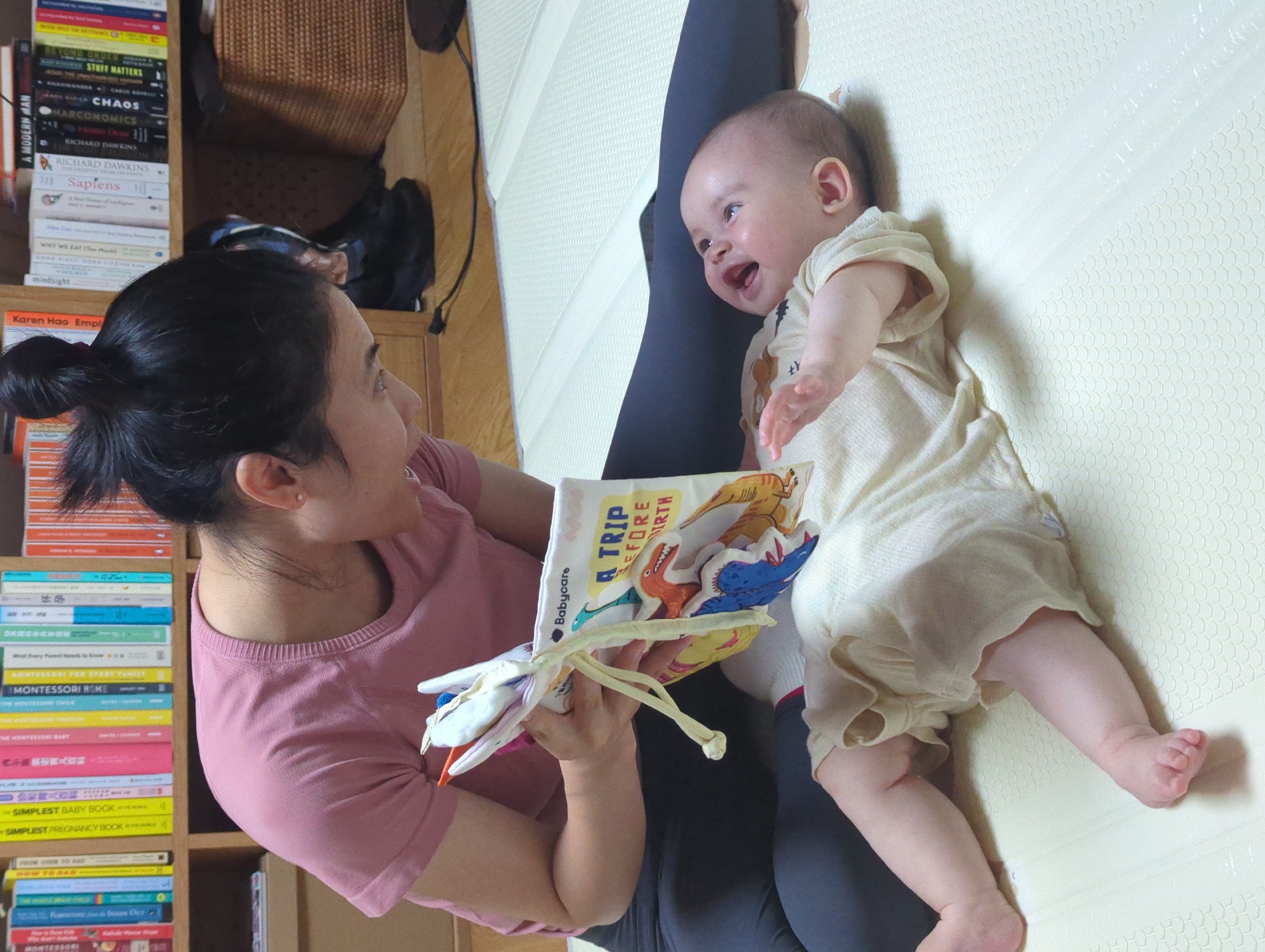A woman with her dark hair in a bun sits cross-legged, facing a baby lying on a light yellow patterned surface. The woman holds a soft book open for the baby to view, the cover titled "A TRIP BEFORE BIRTH". The baby is fair-skinned and laughing, wearing a light-colored romper. The woman is wearing a light pink t-shirt. Behind the woman, a bookshelf is filled with books, including several titles by Richard Dawkins and one titled "Sapiens". Various other books are visible, including Montessori-related books and Chinese encyclopedias. A woven basket and a dark-colored bag are on the shelf above the books. The exact location in the city of Shunde District, China cannot be determined from the provided image.
