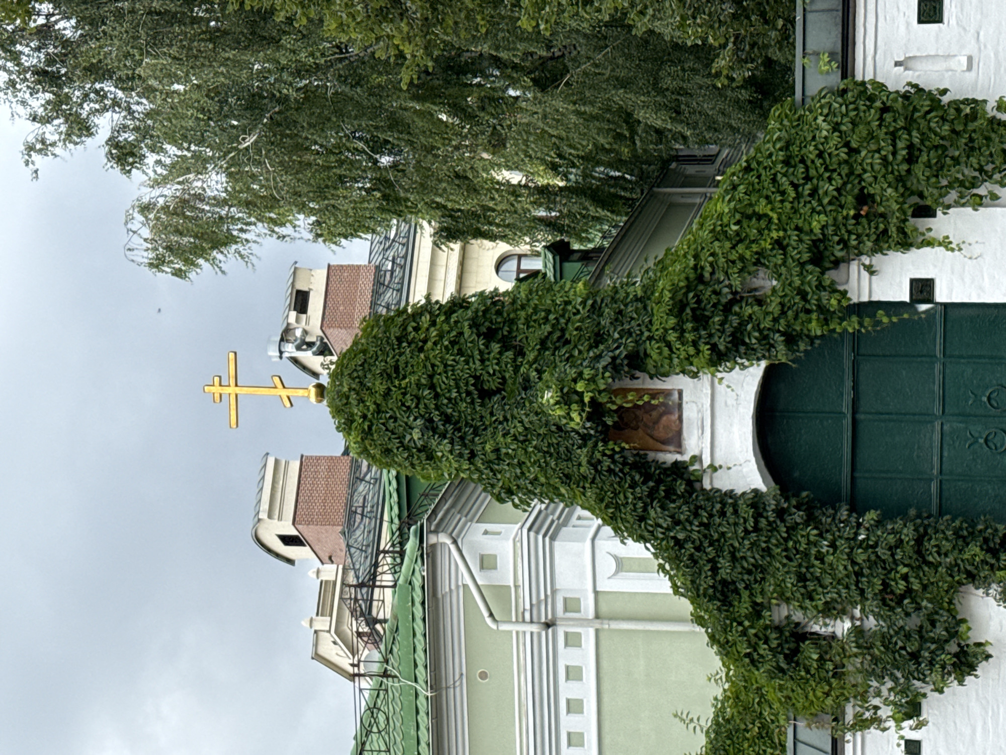 This image captures the entrance to the Church of the Holy Trinity in Ostankino, Moscow, Russia. A large, ornate golden Orthodox cross crowns the structure, which is heavily draped in lush green ivy, obscuring much of the architectural details. Below the cross, a small icon is visible, depicting a religious figure. The entrance itself is a large, dark green double door. The surrounding buildings feature light green and white facades, with green roofs. The environment appears to be an overcast day, with a muted, grey sky. Trees, predominantly birch trees with their characteristic white bark, are visible to the right, their foliage creating a dense canopy. The scene is serene and visually dominated by the contrast between the vibrant green of the ivy and the weathered stone of the buildings.