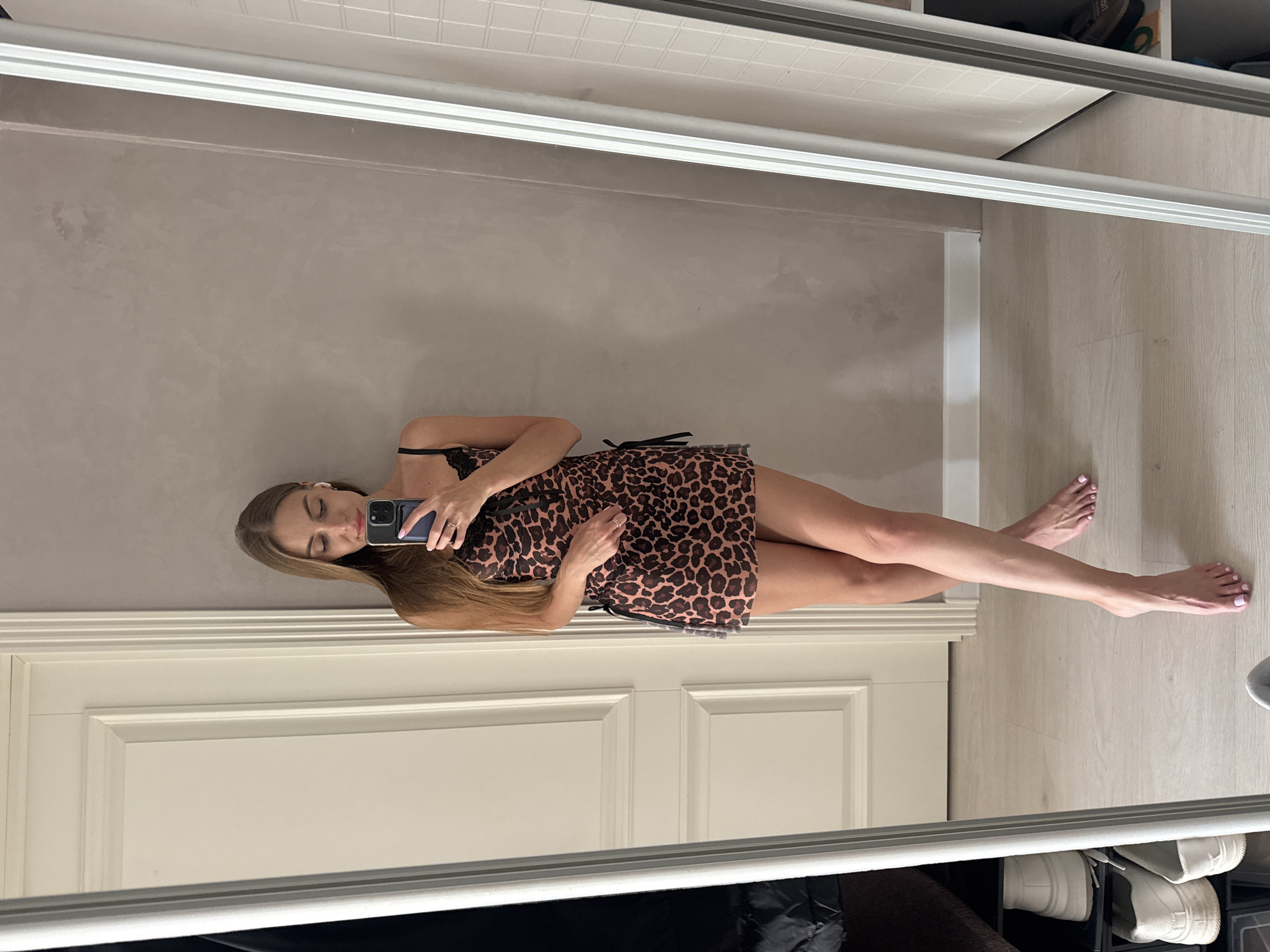 A young woman with long brown hair is standing in a room, taking a mirror selfie. She is wearing a short, leopard-print slip dress with black lace trim. Her left leg is bent and crossed over her right leg. She is holding a smartphone in her right hand, pointing it at the mirror. The room appears to be a bedroom or dressing area, with a white door and a beige wall visible behind her. A large mirror is positioned to her left, reflecting her image and part of the room. In the reflection, a shoe rack with white sneakers can be seen on the left. The floor is light-colored laminate.