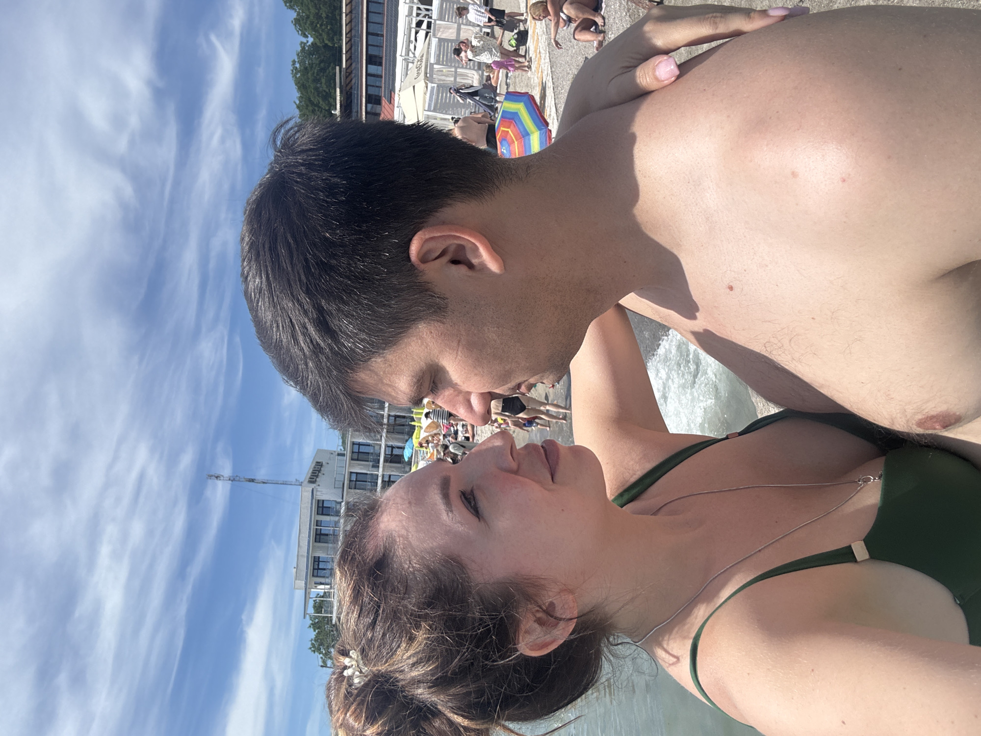 The image shows a close-up of a couple on a beach, with their faces turned towards each other. The woman is on the left, with her hair up in a bun and wearing a green bikini top. The man is on the right, with dark hair and bare chest. They appear to be in an intimate moment. In the background, there are other beachgoers, umbrellas, and buildings, suggesting a lively beach environment. The sky is blue with scattered clouds. The location is identified as Odesa, Ukraine.