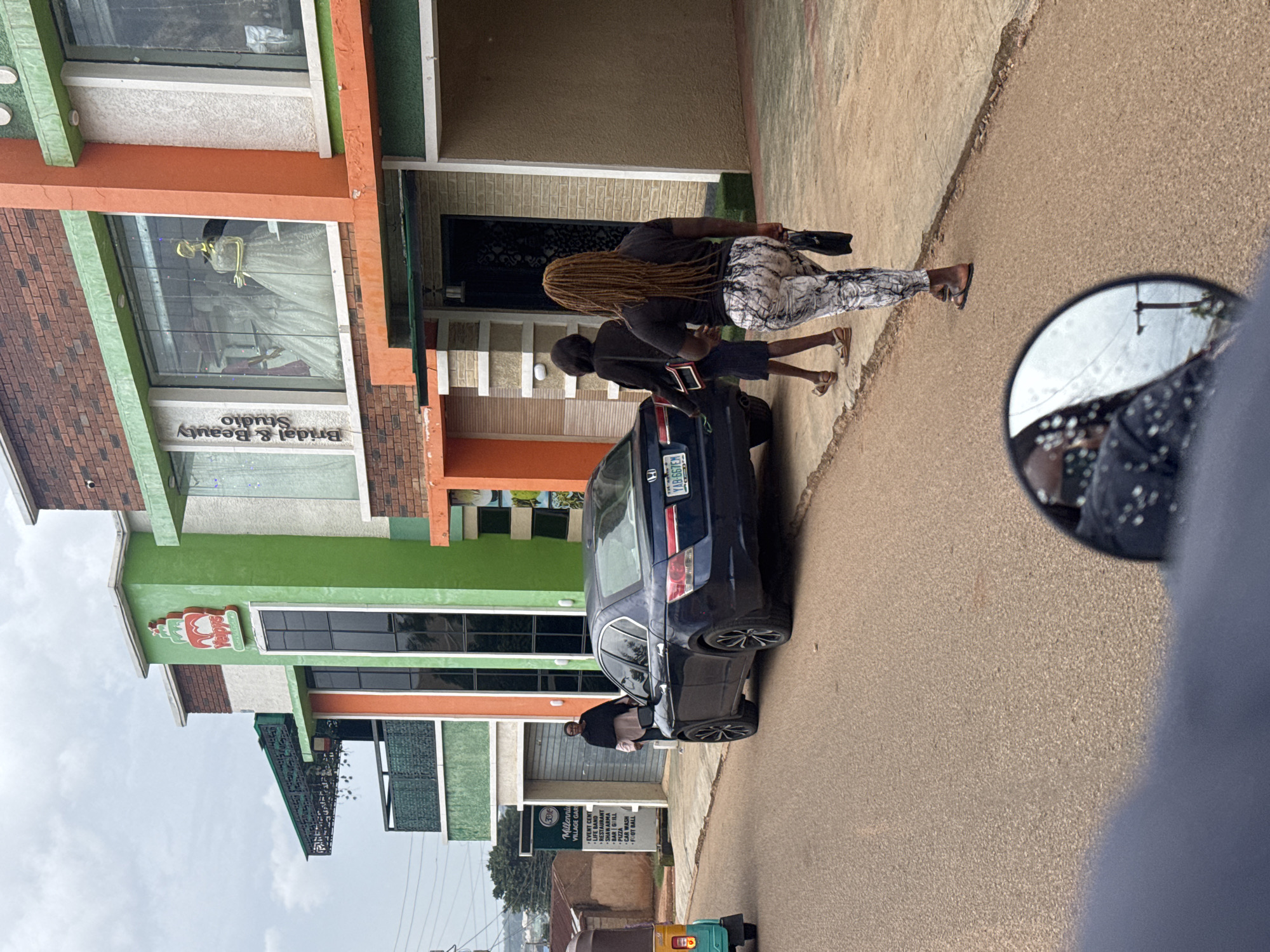 This image depicts a street scene in Gura Top, Nigeria. The primary subjects are two individuals, one with long, blonde braided hair wearing patterned leggings and a dark top, and another person in a dark top and shorts. They appear to be walking along a paved road next to a building. A dark blue sedan is parked on the side of the road. The building features an orange and green facade with large windows. One window displays a mannequin in a wedding dress, and the text "Bridal & Beauty Studio" is visible below it. Another part of the building has a colorful sign with the text "30", "Millenniu", "VILLAGE GAR", and a list of services including "EVENT CENT", "RESTAURANT", "SHAWARMA", "BAR GRILL", "PIZZA", "CAR WASH", and "FOOT BALL". The sky is overcast, suggesting cloudy weather. A motorcycle with a covered passenger is also visible further down the road. A motorcycle mirror reflects part of the scene, including the sky and the person taking the photo. The overall scene appears to be during daylight hours.