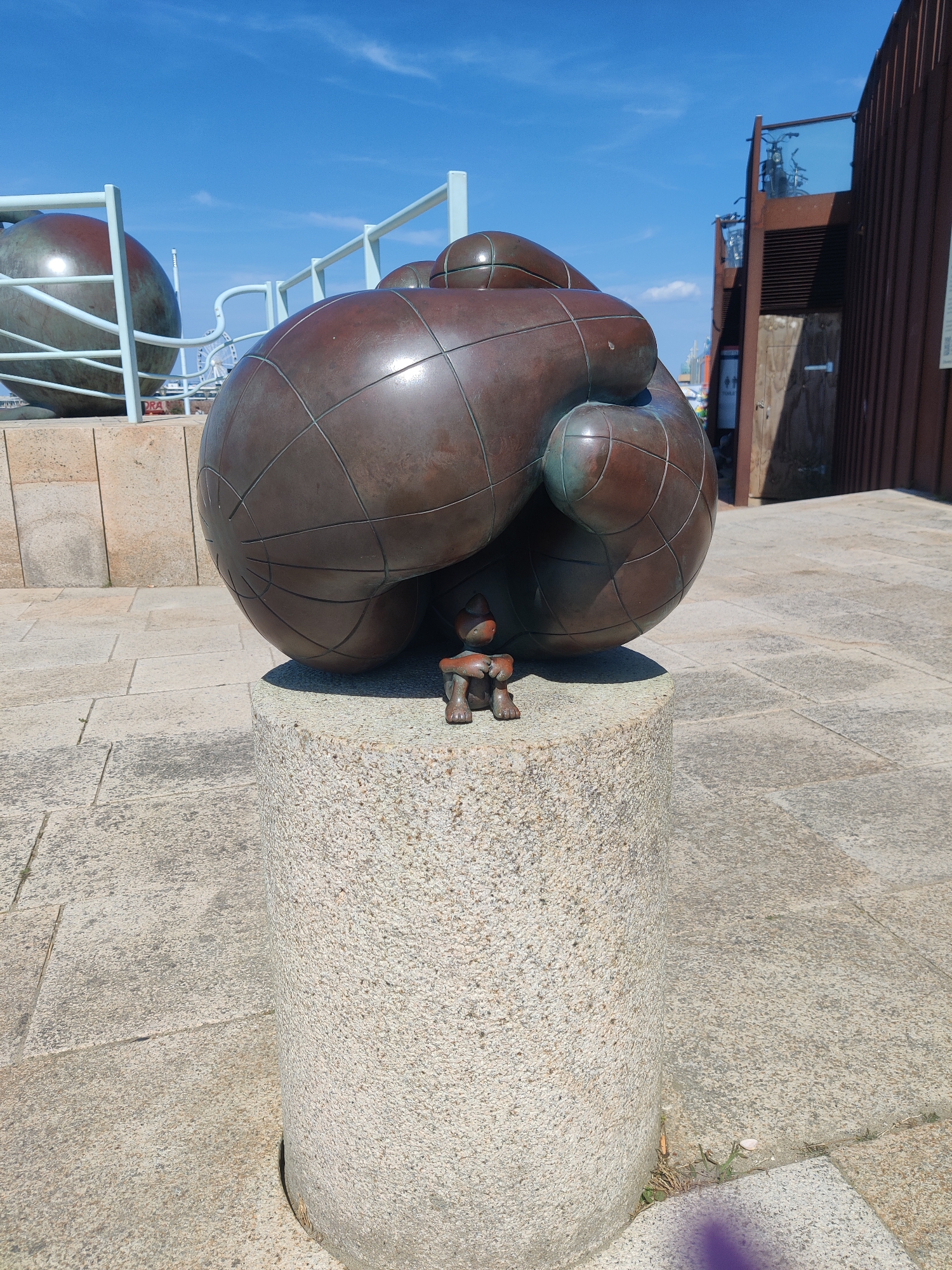 A bronze sculpture of a squatting figure is positioned on top of a rough, granite cylindrical plinth in front of a larger, dark bronze spherical sculpture marked with a grid pattern. The larger sculpture appears to be a stylized globe or planet, with bulbous sections. In the background, there are other spherical sculptures on metal stands, a fence, a brown building with glass, and a blue sky with scattered white clouds. The ground is paved with square, light-colored tiles. The scene appears to be outdoors in a public space. This specific sculpture is located at the "Beeldend Monument" or "Visual Monument" by artist Henk Visser, situated near the Zuiderpark in The Hague, Netherlands.
