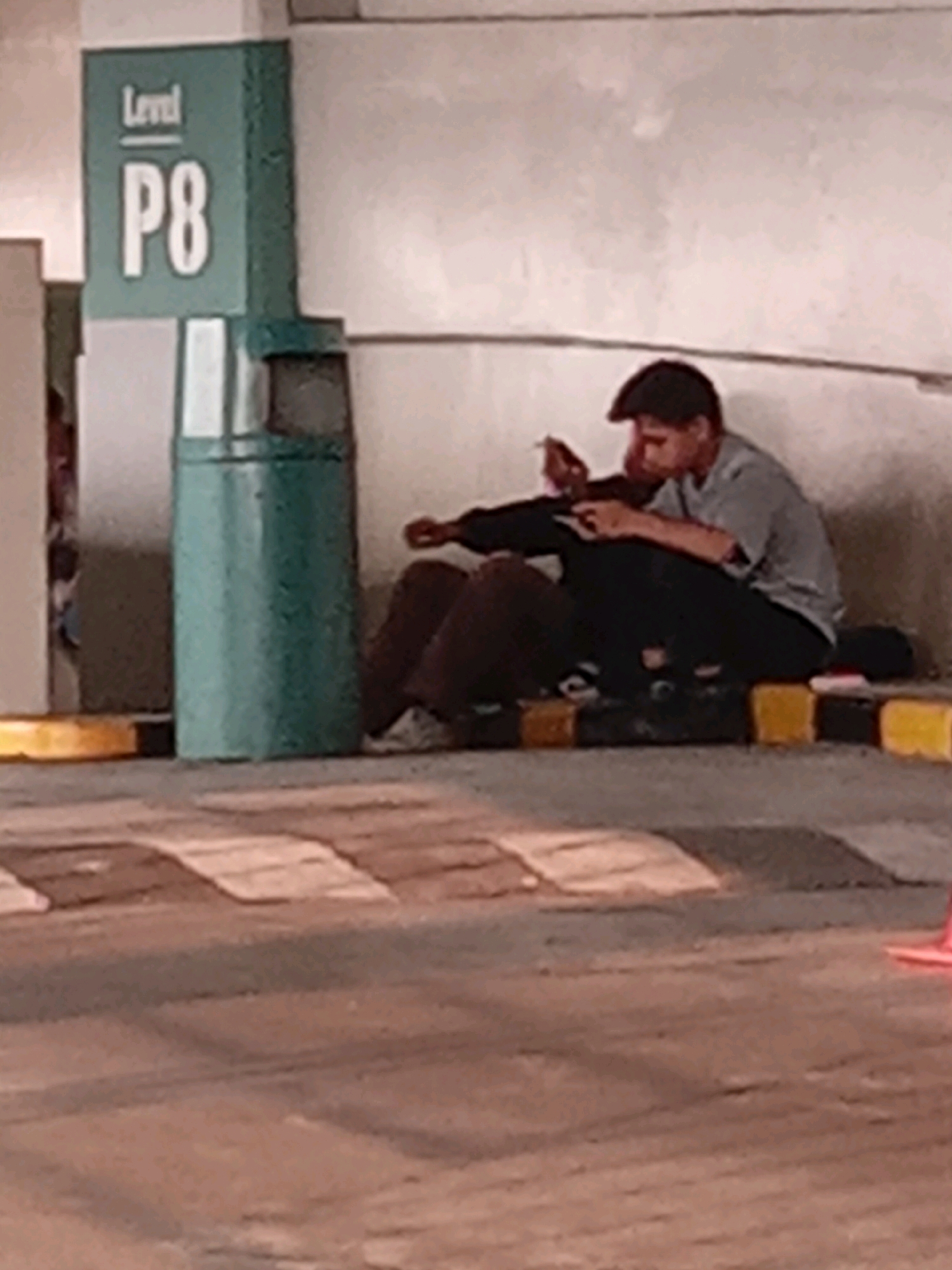 In an indoor parking garage, two individuals are seated on the ground next to a teal trash receptacle and a pillar marked "Level P8". The person on the left is wearing a brown shirt and dark pants, while the person on the right is wearing a grey t-shirt and dark pants, with a flat cap on their head. The individual on the right appears to be holding a cigarette and a phone. The flooring is a mix of asphalt and tiled areas, with yellow and black striped parking bumperettes visible. The overall lighting suggests a dimly lit environment, typical of a parking structure. The scene is likely located within the city of Surabaya, Indonesia, given the context provided.