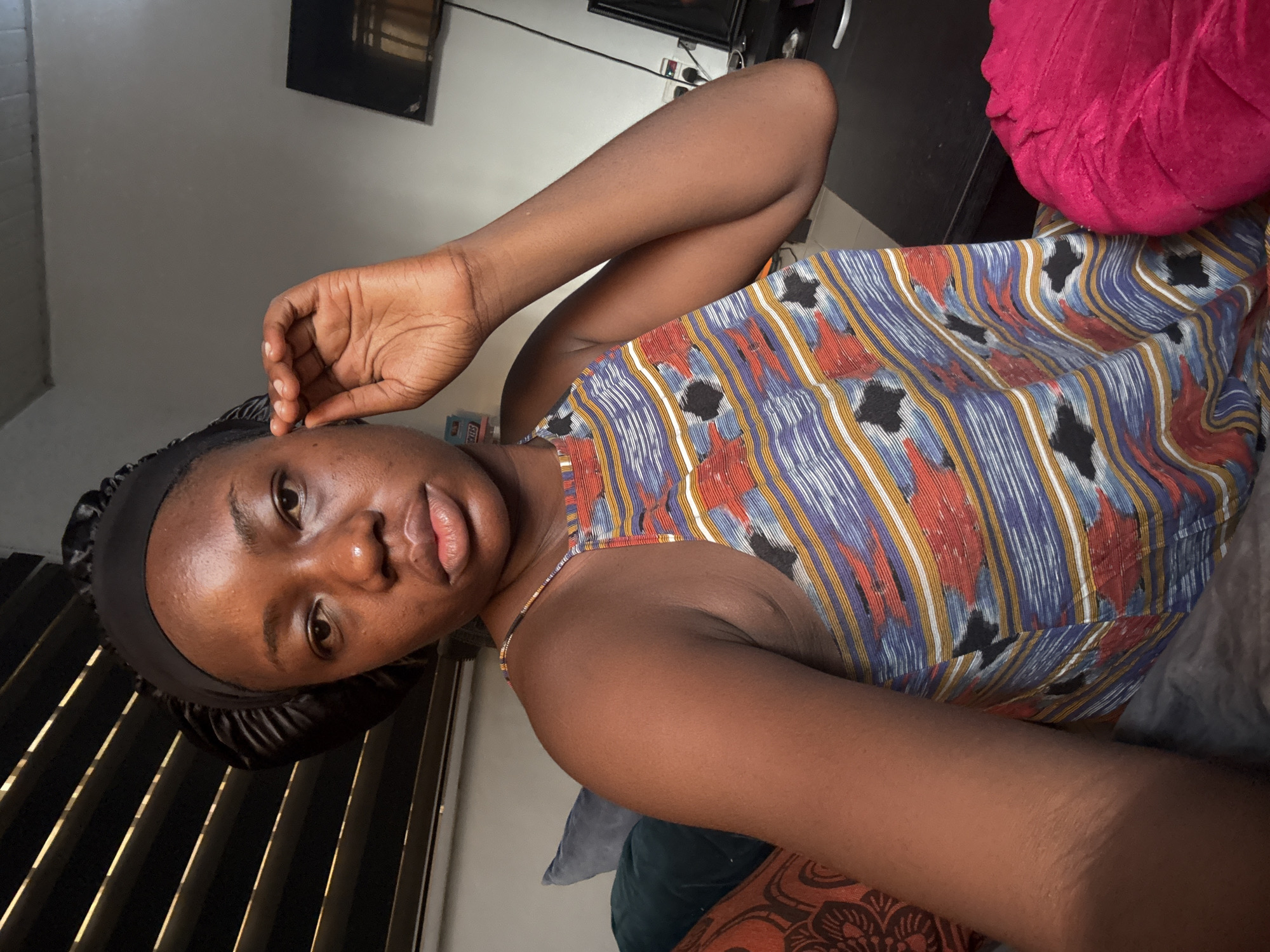 A young woman is captured in a close-up selfie, reclining indoors. She has a medium-dark complexion, dark eyes, and full lips with a subtle sheen, bearing a relaxed expression with a slight pout. Her dark hair is covered by a shiny black satin-like headwrap or bonnet. She is wearing a camisole-style top featuring a vibrant, multi-colored geometric pattern of blue, white, yellow, red, and black stripes and abstract shapes. Her right hand is raised, with fingers gently touching her forehead.
She appears to be seated or lying on a bed or sofa. Next to her head, a bright fuchsia-pink, textured pillow is visible. Another patterned cushion or blanket is partially seen. The background shows vertical window blinds on the left, suggesting a window, and dark furniture, possibly a desk or media console, on the right. A television screen is discernible on the furniture. White electrical outlets or switches are mounted on the light-colored wall.
The activity is a self-portrait taken from a slightly high angle. Visible text on small items in the background includes "ASTRO" and "EXTRA." The setting is an indoor space, with the location context provided as Abuja, Nigeria.
