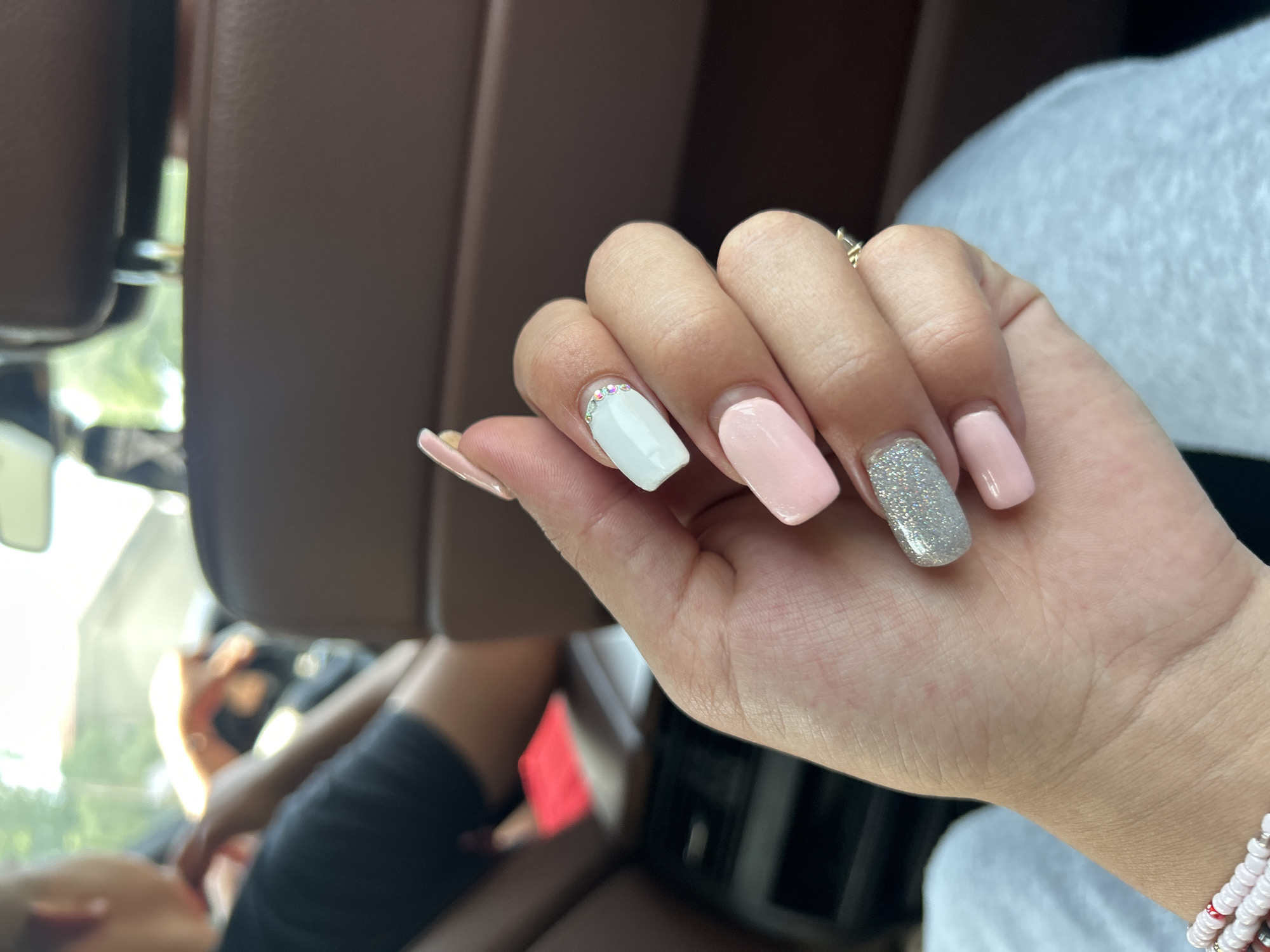 The image presents a close-up view of a person's right hand, showcasing a fresh manicure. The nails are a medium length with a square or squoval shape. The color scheme is varied, featuring pale pink on the thumb and pinky finger, a light blue or mint green on the index finger, and a sparkling silver glitter on the middle finger. The light blue nail is adorned with a line of small, clear rhinestones or gems placed along the cuticle. A gold-toned ring is visible on one of the fingers. On the wrist, a multi-strand beaded bracelet, primarily with white/pink and some red beads, is worn.
The background is out of focus, suggesting a shallow depth of field. It appears to be the interior of a vehicle, likely a car, with soft brown upholstery visible. Bright daylight streams in from the left side of the frame, illuminating blurry green foliage visible through a window. Partially visible in the background, out of focus, are shapes that suggest another person or parts of the vehicle's interior. The setting indicates that the photo was taken during the daytime, likely on a sunny day given the bright light. There is no visible text in the image.