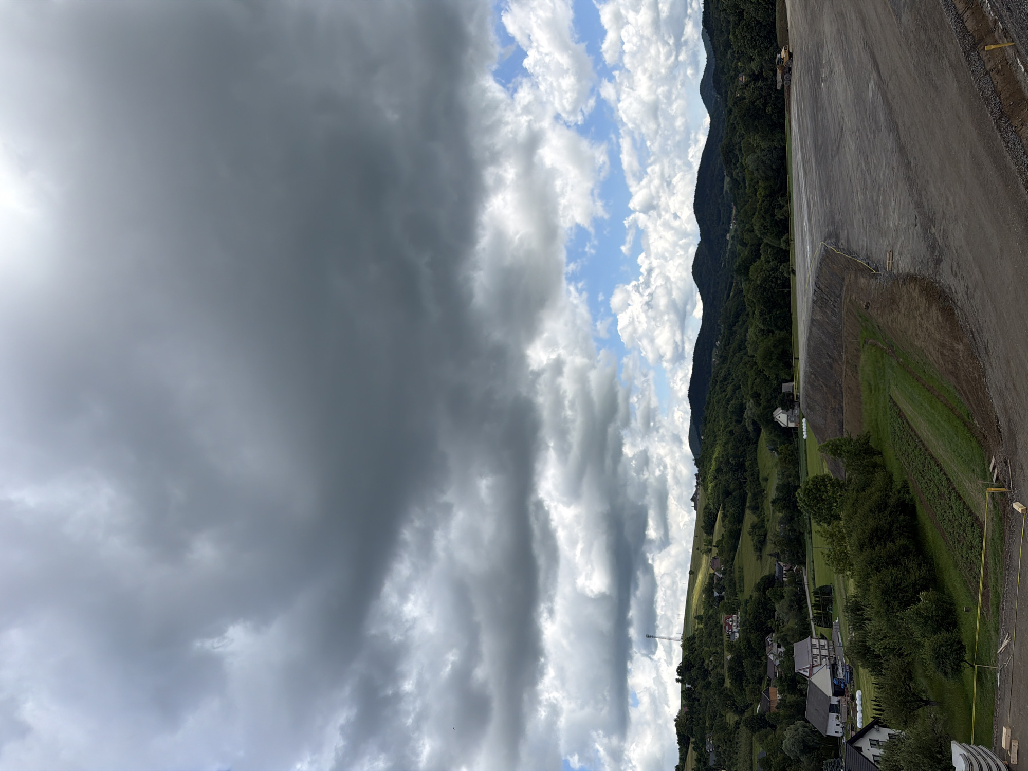 The image shows a construction site with a large, cleared area of dark gravel. In the background, there are rolling green hills dotted with houses and trees. The sky is filled with dramatic, dark clouds, with patches of blue sky and white, fluffy clouds peeking through. A yellow construction barrier is visible along the edge of the cleared area. Further in the background, a yellow construction vehicle, possibly a dump truck, is parked. The landscape suggests a rural or semi-rural setting, possibly in Stróża, Poland, given the context. The overall impression is one of an overcast day with a construction project underway in a scenic, hilly environment.