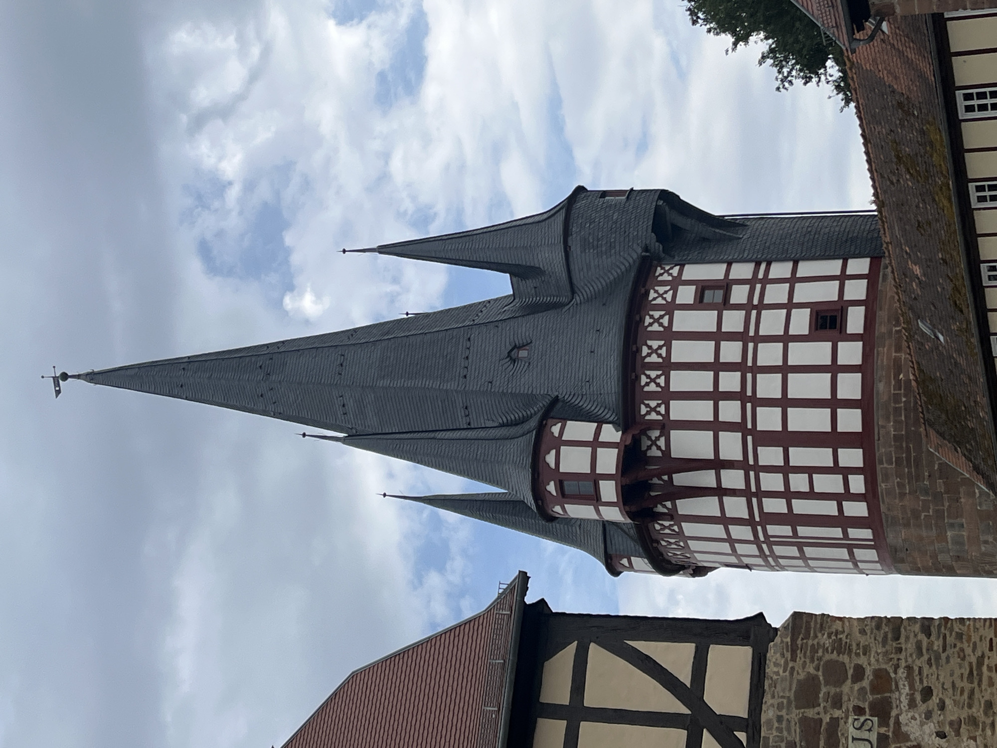 The image depicts a close-up, vertically oriented view of a historic building with a prominent, pointed spire, characteristic of medieval architecture. The spire is covered in dark gray slate tiles, and it tapers to a sharp point. Below the spire, the building features a section with a distinctive half-timbered design, characterized by white infill panels and dark brown wooden beams arranged in a grid pattern. Some of these beams form decorative "X" shapes. Small, dark windows are visible within the white panels.
The building's construction also includes red brickwork. Adjacent to this structure, another building with a red tiled roof and a stone facade is partially visible. The sky above is filled with scattered white clouds against a pale blue background, suggesting it is daytime with variable weather.
The scene appears to be a tourist attraction or a historical site, possibly a castle or a well-preserved old town area, given the architectural style. There is no visible activity or people in the frame. The lighting suggests it is likely mid-day. No text is clearly legible in the image. The location context provided is Neustadt, Germany, which aligns with the style of architecture often found in that region.