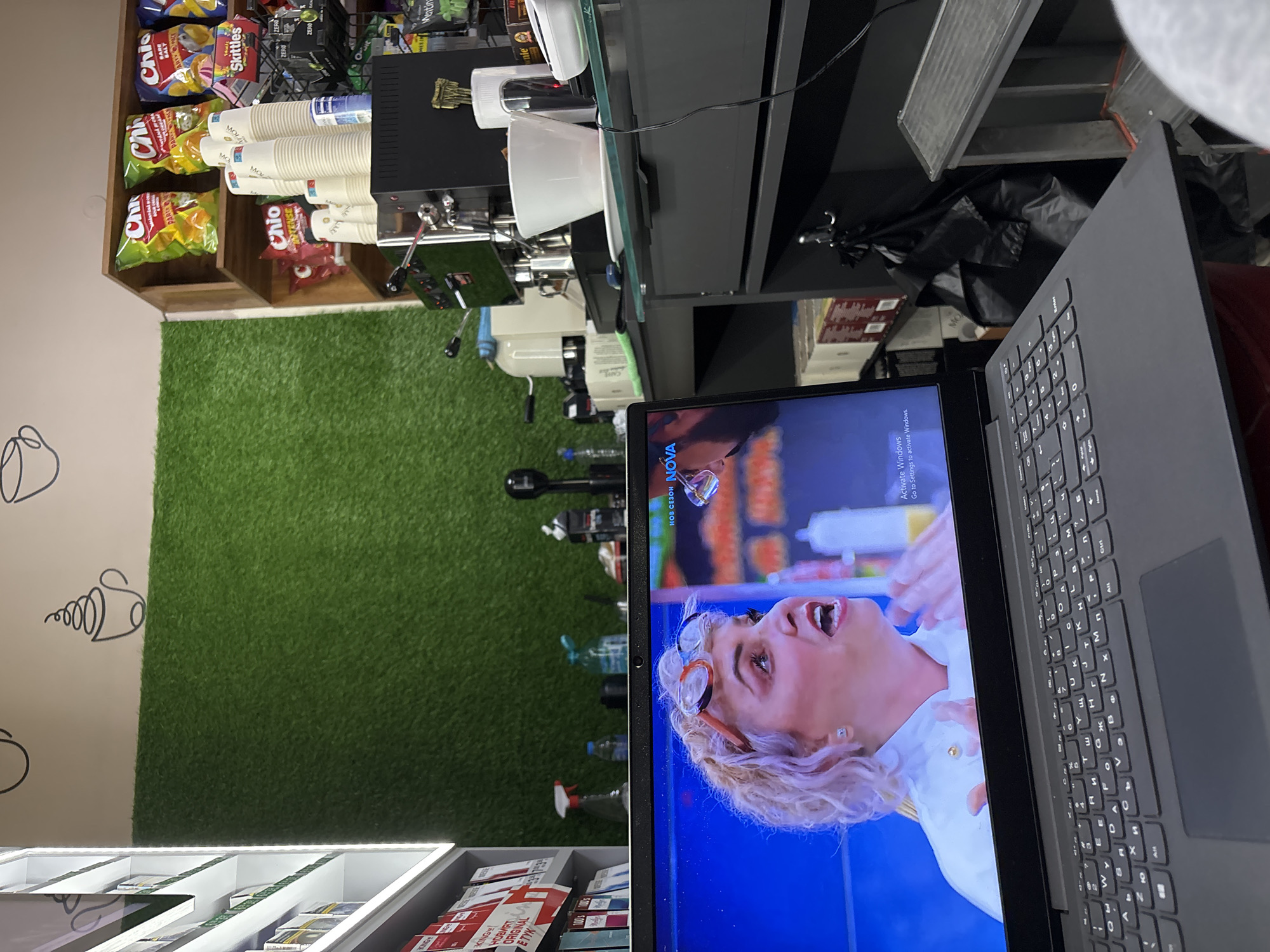 The image presents a skewed view of a shop interior, possibly located in Plovdiv, Bulgaria, based on the Cyrillic keyboard on a laptop in the foreground and the presence of Bulgarian product packaging. The laptop's screen displays a woman, possibly a chef, from a TV show. Next to the laptop, various items are displayed, including snacks like "Chio" chips and "Skittles" candies. Disposable coffee cups are stacked on a wooden shelf behind the snacks. A coffee machine and various cleaning supplies are also visible. The background includes a wall with coffee-related drawings and a section of artificial grass. The overall environment suggests a small retail space, perhaps a cafe or convenience store.