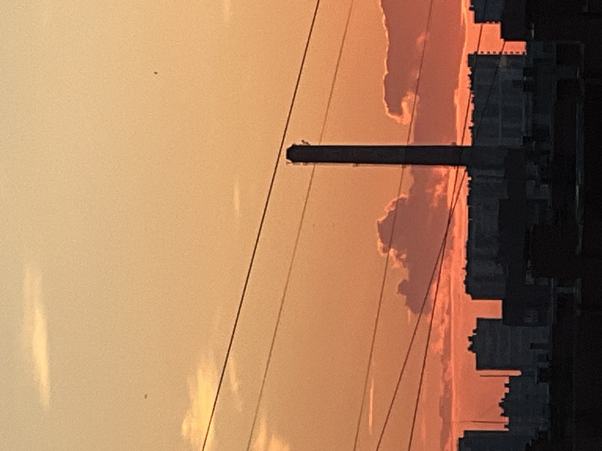 The image depicts a city skyline at dusk or dawn, with a predominant orange and yellow hue across the sky. In the foreground, silhouetted buildings form a dark band across the bottom of the frame. Above these, a tall, slender industrial smokestack stands prominently in the center of the image. Several power lines crisscross the sky. Fluffy clouds, tinged with the warm colors of the sunset or sunrise, are visible behind the smokestack and the buildings. The scene appears to be an urban or industrial area, possibly near Ladozhskoe Ozero, Russia, given the context. There are no people or discernible actions taking place in the image.