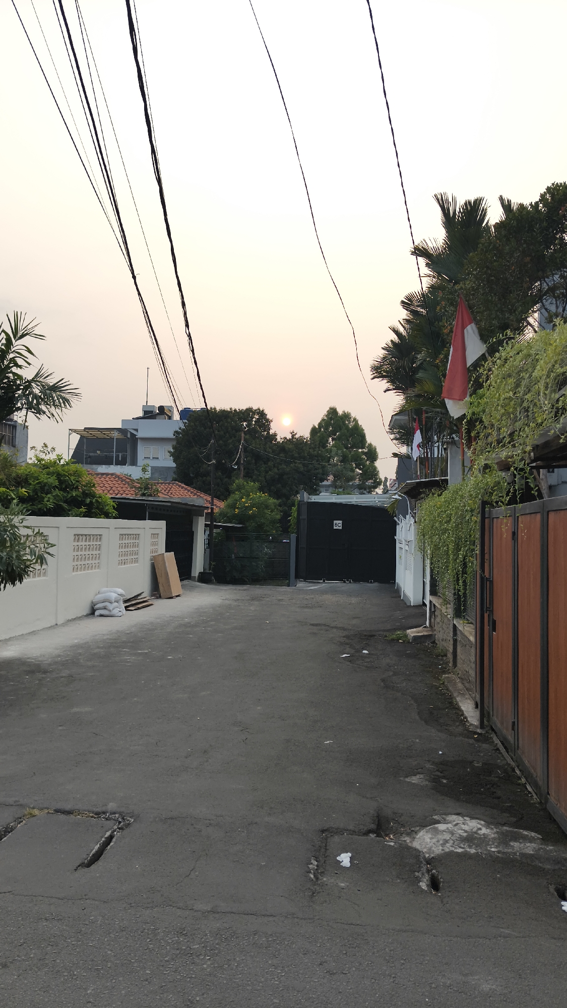 The image depicts a residential street in South Jakarta, Indonesia, during what appears to be sunset. The sky is a pale orange and yellow, with the sun visible as a small, bright circle near the horizon. Overhead, several dark electrical wires crisscross the sky.
On the left side of the street, a white concrete wall with a decorative pattern runs along the property line. Behind the wall, a building with a red-tiled roof and white walls is visible. Piles of white sacks and some wooden planks are stacked near the wall. A darker, open doorway is set into the wall further down the street.
The right side of the street features a tall, dark metal gate, possibly an entrance to a property. A small white square with the letters "BC" is affixed to the gate. Above the gate, palm fronds and lush green foliage are visible, along with an Indonesian flag (red and white) flying. A dense cascade of green vines hangs down from an overhead structure. A wooden fence with vertical planks forms the boundary of another property.
The street itself is paved with asphalt, showing signs of wear and tear, including cracks and two visible manhole covers. Scattered pieces of white litter are present on the road surface. The overall impression is a quiet, suburban street scene at dusk.