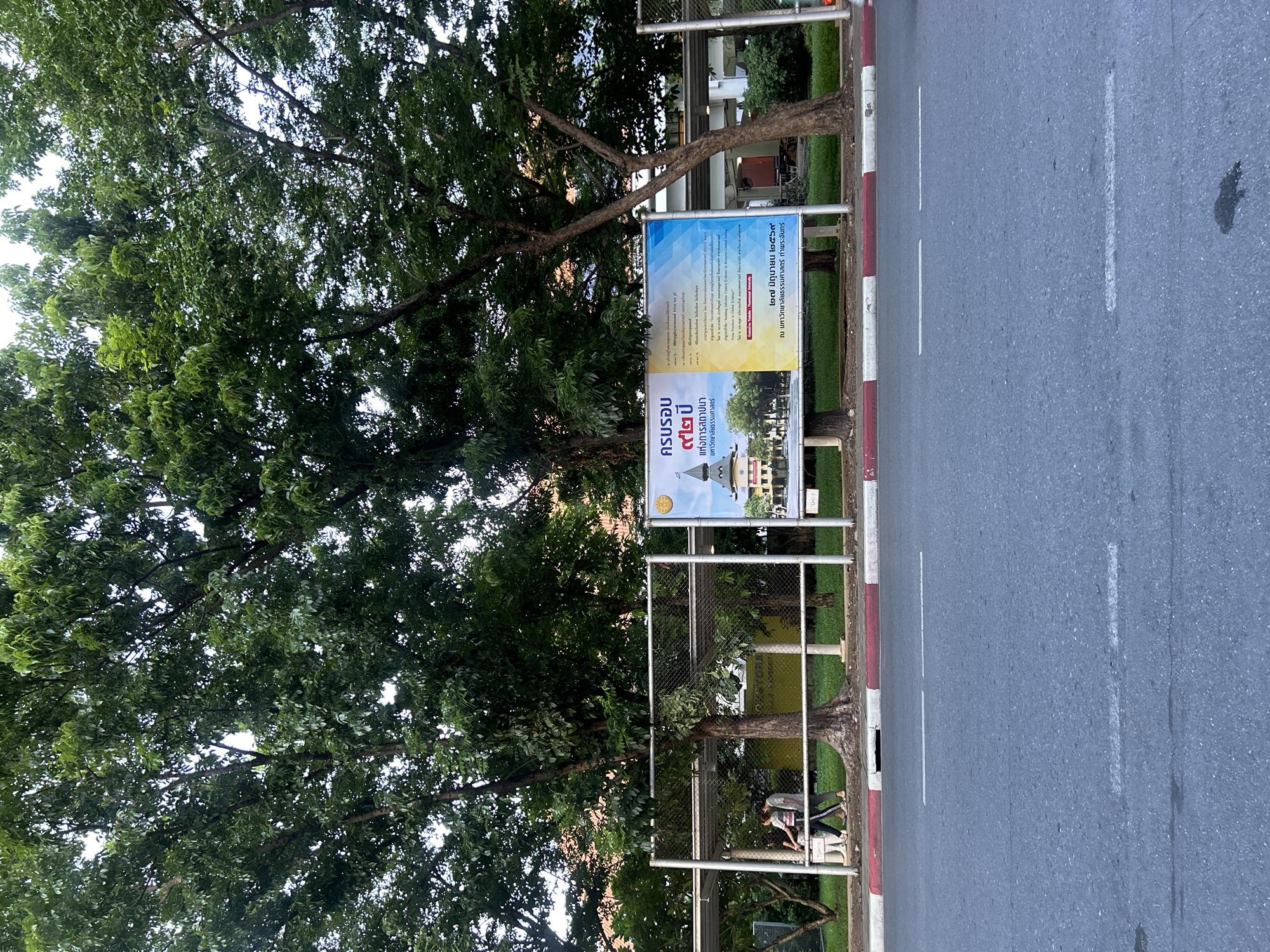 In Tha Khlong Town Municipality, Thailand, a billboard is displayed in front of lush green trees and a street. The billboard features Thai text and images of a university building. Two individuals, a woman and a child, are walking past the billboard. The scene is captured from a street-level perspective, with the road and sidewalk in the foreground. The environment appears to be an outdoor public space, possibly near a university campus.