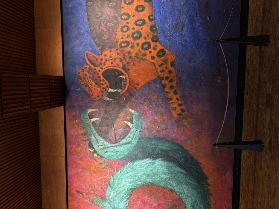 A large mural titled "Duality" by Rufino Tamayo is prominently displayed, depicting a confrontation between two stylized creatures. On the left, a green, feathered serpent with an open, fanged mouth is shown against a mottled red and purple background. On the right, an orange, spotted jaguar-like figure with an open, fanged mouth and an extended clawed paw faces the serpent, set against a deep blue background. The mural is located within the National Museum of Anthropology in Chapultepec Park, Mexico City, Mexico. In the foreground, a polished wooden floor is visible. Two black, conical stanchions with a light-colored rope barrier are positioned in front of the mural. The ceiling above the mural consists of parallel wooden slats.