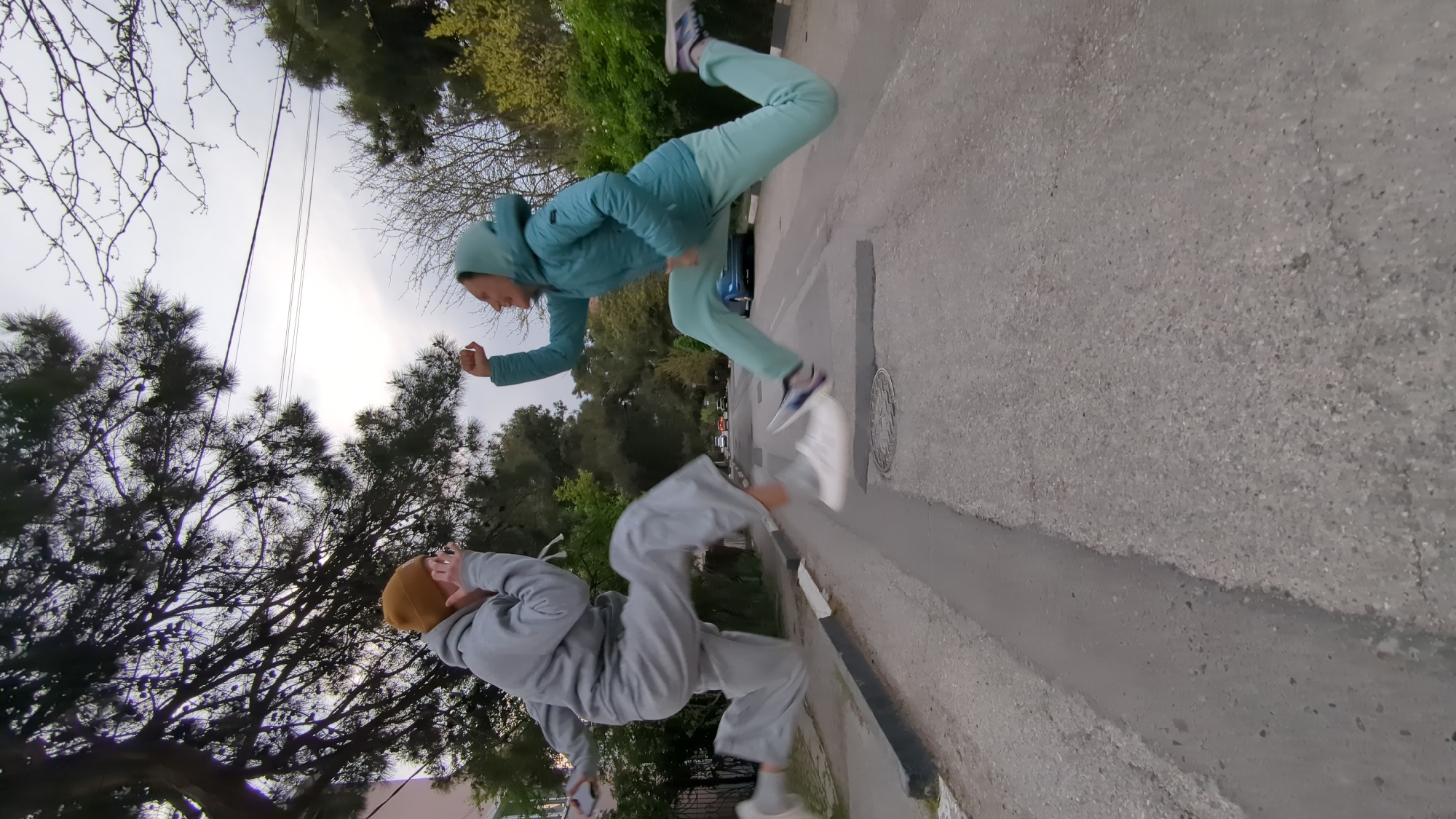 This image depicts two individuals engaged in dynamic activity on a paved street in Gelendzhik, Russia. The photograph is taken from an unusual, rotated perspective, making the ground and curb appear as a vertical surface.
One person, positioned towards the top of the frame (when viewed upright), is wearing a teal or mint green hooded tracksuit and sneakers. They are captured mid-air, with knees bent and arms raised, suggesting they are performing a jump or a parkour-style maneuver. Their body is angled dynamically, and their facial expression, though slightly blurred, conveys focus on the action.
The second person, located towards the bottom of the frame, is dressed in a grey hooded tracksuit and a mustard yellow beanie. They are also in a vigorous pose, with bent legs and arms raised near their head, indicating they are in the midst of a jump, crouch, or other acrobatic movement. Their face is partly obscured by their hands or the angle of their head.
The setting is an outdoor urban or suburban environment. The paved surface features a distinct curb and a gutter running alongside it. A circular manhole cover is visible on the pavement. In the background, various trees, including both deciduous trees with bare branches and evergreen pines, are silhouetted against an overcast sky. Overhead utility wires can also be seen. Distant cars are faintly visible further down the street. The lighting is diffused, characteristic of an overcast day, possibly late afternoon or early morning, with no strong shadows. There is no visible text in the image.