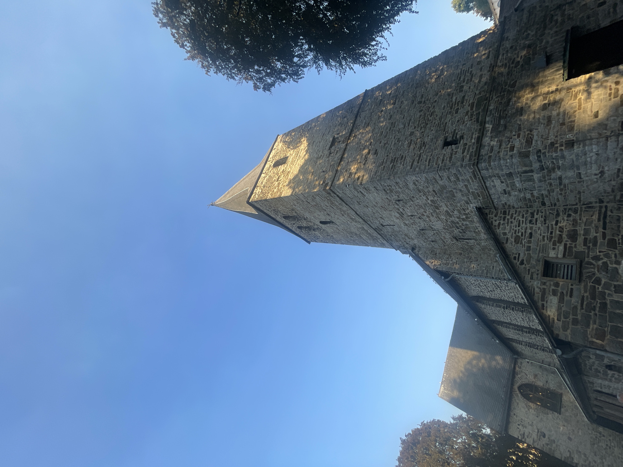 An upward-angle, medium shot captures the stone tower and adjacent building of the Protestant church in Iserlohn, Germany, under a clear blue sky. The tower, made of rough-hewn stone blocks, has a pointed steeple topped with a cross. Small, rectangular windows dot the tower's facade, and shadows from nearby trees cast a dappled pattern on its surface. The adjacent building, also stone, has a pitched roof covered with dark shingles. One gothic-style arched window is visible, along with smaller rectangular windows. Trees frame the right side of the tower and the left side of the adjacent building, their foliage dark and dense. The lighting suggests it is either early morning or late afternoon.