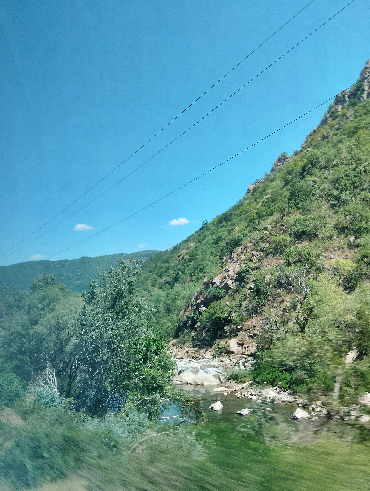 The image depicts a scenic view of a river flowing through a mountainous, forested area. In the foreground, the water is a vibrant green, reflecting the surrounding vegetation. Several light-colored rocks are visible in and around the river. The left side of the image features lush green trees, some of which appear blurred due to motion. To the right, a steep, rocky mountain slope is covered with dense green foliage and scattered rocks. The sky above is a clear, bright blue with a few wispy white clouds. Three thin, dark power lines stretch diagonally across the upper portion of the sky. In the background, rolling hills covered in greenery recede into the distance. The overall impression is one of a natural, tranquil landscape, likely captured from a moving vehicle given the motion blur in the foliage. The location is identified as being in the vicinity of Stara Kresna, Bulgaria.