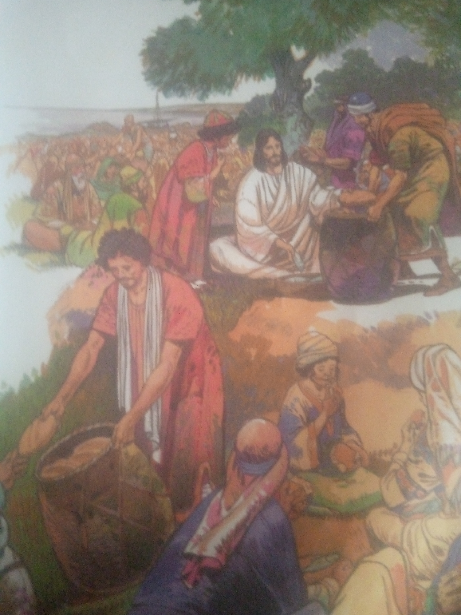 The image depicts a biblical scene, likely the feeding of the multitude, set outdoors with a large gathering of people. In the foreground, a person in an orange robe with a white scarf draped around their neck is bent over a large woven basket containing loaves of bread. To their right, another figure in a purple robe and blue headdress is seated. In the middle ground, a man with a beard, dressed in white, is seated and appears to be receiving or interacting with a loaf of bread. Other individuals are visible in the background, some seated, some standing, in various colored robes. The environment includes a body of water in the distance, green foliage, and a large tree with a spreading canopy. The overall atmosphere suggests a communal gathering or event. The location in Jauro Yinu, Nigeria is not discernible from the image itself.