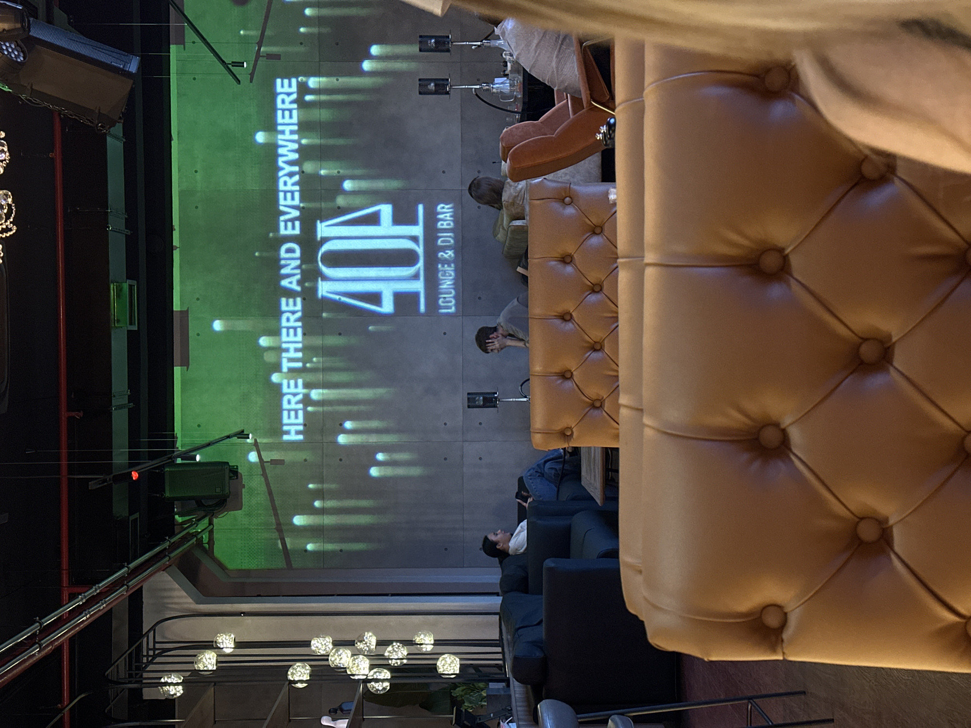 The image captures the interior of a contemporary lounge or bar, likely in the evening, viewed from an elevated vantage point. The setting features a blend of modern industrial design and plush comfort.
In the foreground, a prominent, tufted leather sofa in a warm caramel-brown color takes up much of the right side, showing intricate button detailing. Below and further back, darker-colored sofas and armchairs are arranged, providing seating for patrons. Several individuals are present, appearing relaxed; one person is visible on a dark sofa looking up, and another is seated on a brown sofa.
A large projection dominates the background wall, displaying dynamic, glowing vertical lines against a greenish hue. Bold text on the projection reads "HERE THERE AND EVERYWHERE". Below this, the name of the establishment, "404", is prominently displayed in a stylized font, followed by the description "LOUNGE & DJ BAR".
Overhead, an industrial aesthetic is evident with exposed red pipes and various lighting fixtures, including a large rectangular light source and smaller directional lights. Decorative lighting fixtures featuring clusters of glowing, spherical elements hang from the ceiling in the mid-ground. The overall activity depicted is people socializing and relaxing in a modern entertainment venue in Vladimir, Russia.