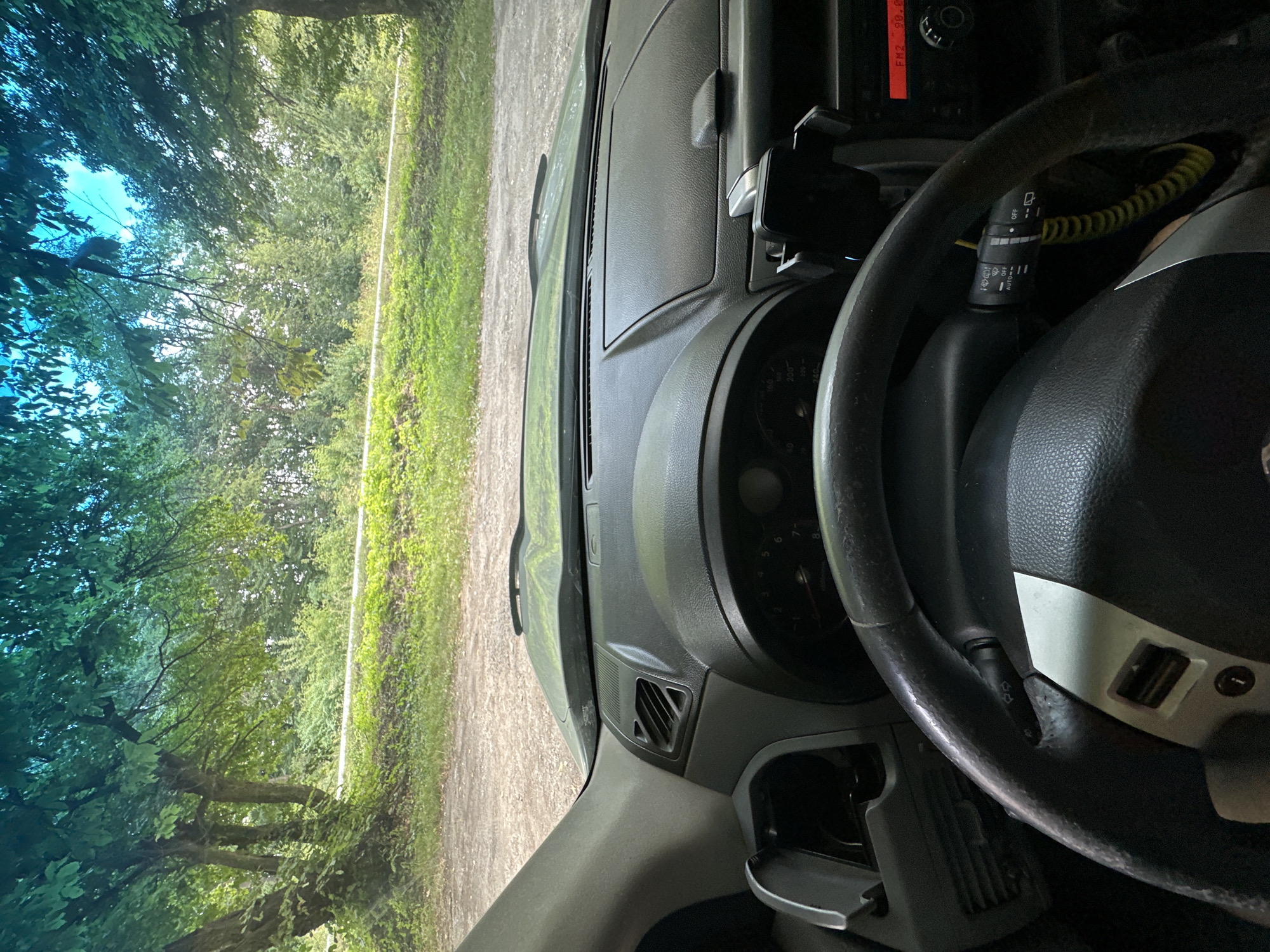 A first-person perspective from inside a car shows a view through the windshield of a dirt road lined with lush green trees and vegetation. The dashboard and steering wheel are visible in the foreground, with the car parked facing a wooded area. The sky is partially visible through the trees and appears to be overcast with blue patches. The scene depicts a natural, outdoor setting.