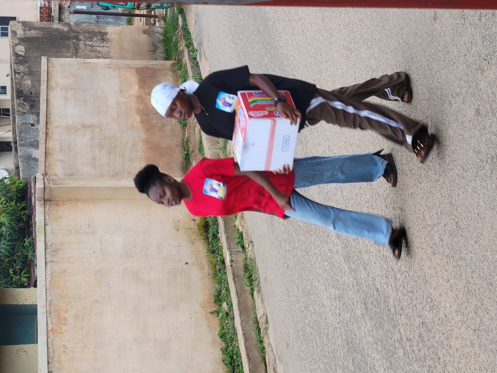 In Jos, Nigeria, two young women walk along a paved street. The woman on the left, wearing a red t-shirt and blue jeans, walks with her head turned to the left. The woman on the right, in a black t-shirt and brown track pants, carries a white and red box. Both women are wearing sandals. The background features a textured wall and some vegetation, suggesting an outdoor urban setting. The lighting indicates it may be daytime.