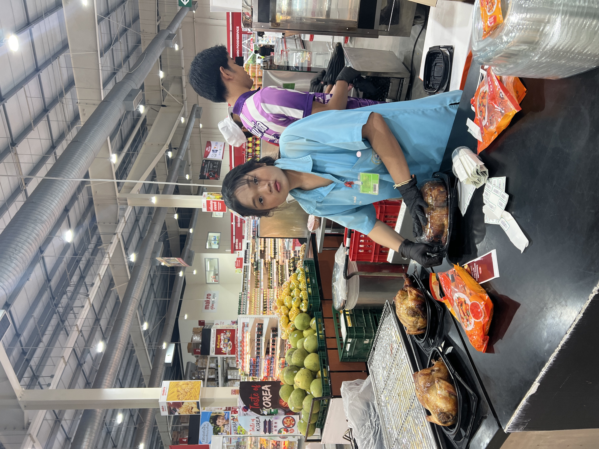 This photograph, taken in what appears to be a Makro store in Baan Hua Thanon, Thailand, shows two individuals working at a checkout counter. A female worker, wearing a light blue uniform, name tag, and black gloves, is facing the camera while handling food items, possibly rotisserie chickens, in black containers. Barcodes and labels are present on the counter near her. Behind her stands another worker, presumably male, wearing a purple and white striped shirt and black gloves. The counter is black and contains various food items, including packaged sauces or spices. In the background, shelves stocked with a variety of products can be seen. A display of mangoes sits near the counter. The store's ceiling is visible, showing overhead lighting and ductwork. Banners and signage are displayed, including the "makro" logo and promotions for Korean food products.