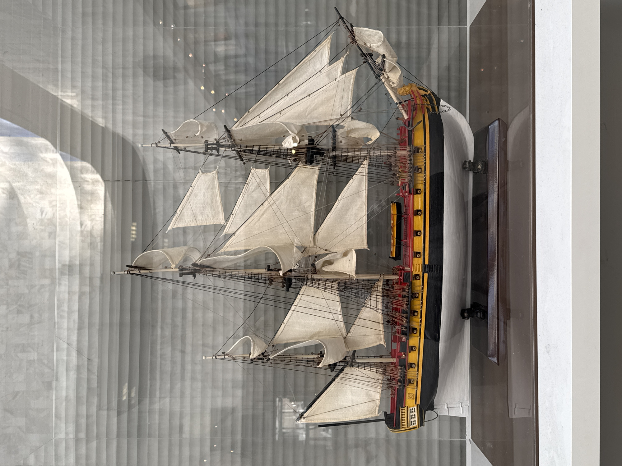 The image shows a model ship in a glass display case. The ship has white sails and a black and yellow hull with red accents. It is a detailed model, with visible rigging and small details. The display case is in a museum setting, with vertical lines reflecting in the glass, suggesting it's against a window. The model ship is likely located in the Central Naval Museum in Saint Petersburg, Russia, which houses a collection of naval models and artifacts.