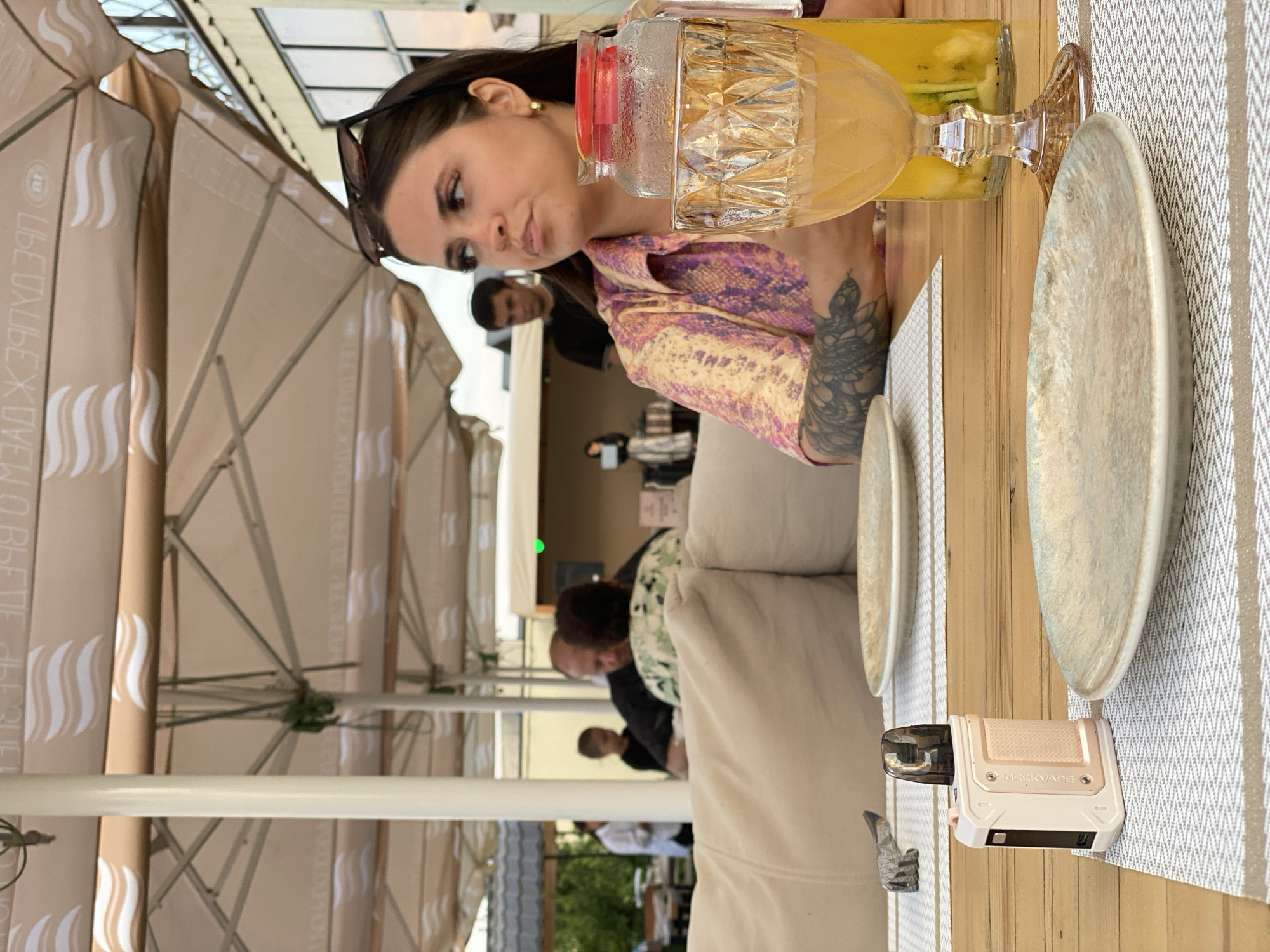 In Derbent, Russia, an outdoor restaurant setting features a woman with dark hair and sunglasses perched on her head, wearing a patterned shirt, seated at a wooden table. In front of her is a glass pitcher filled with a yellow beverage containing fruit, and a decorative glass. On the table, two plates are placed on placemats, and a light-colored vape device is positioned on one of them. In the background, other patrons are visible, including a man and a child, and umbrellas provide shade over the seating area. The overall scene depicts a casual dining experience in an urban environment.