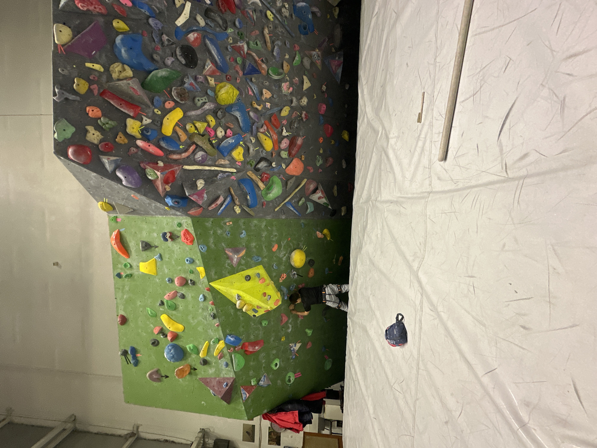 The image shows an indoor climbing gym. A climber is positioned on a green wall with various colored holds. Above the green wall is a gray wall also filled with climbing holds. To the right of the climbing walls is a white wall with what appears to be a hanging bag. A long stick is also visible on the white wall. There is no clear indication of the location as Sofia, Bulgaria.
