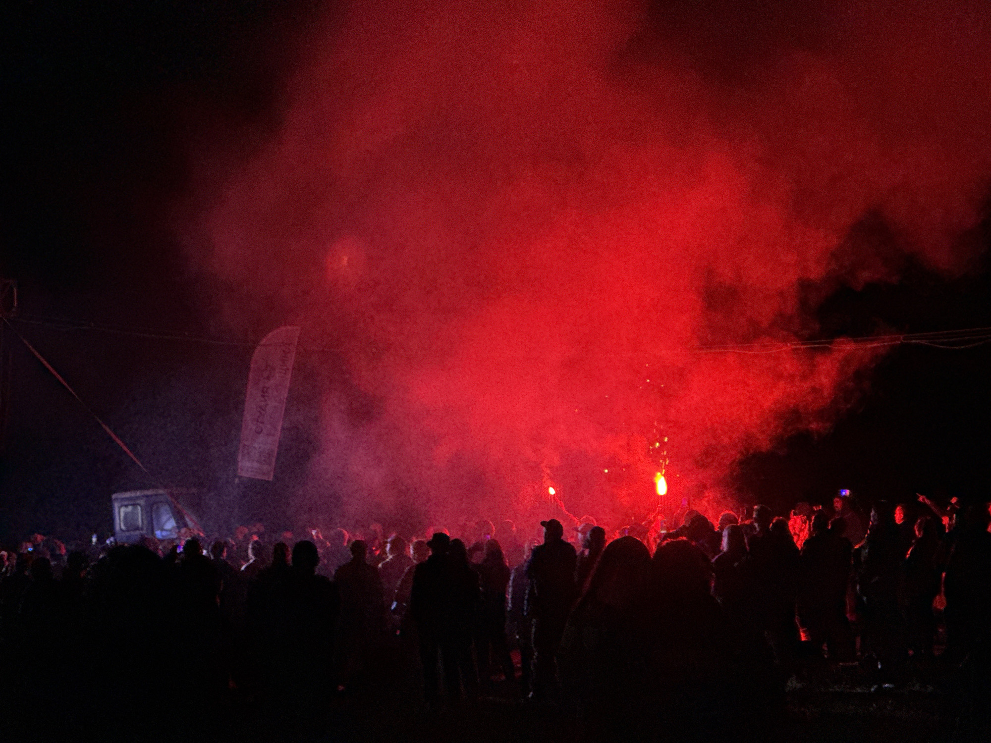 The image captures a nighttime outdoor gathering in Большая Кивара, Russia. A dense crowd of individuals occupies the lower and mid-ground, predominantly appearing as dark silhouettes against a backdrop of intense red light and pervasive smoke or haze. The red illumination emanates primarily from the right side of the scene, where multiple individuals within the crowd are observed holding and emitting bright red flares or pyrotechnic devices, which contribute to both the light and the smoke. In the upper-left quadrant, a dimly lit, box-shaped vehicle, possibly a utility van or small truck, is present. Above this vehicle, a vertical white banner is visible, displaying partially discernible text, which appears to be Cyrillic. Multiple dark wires or cables stretch across the upper-left portion of the frame. The ground beneath the crowd is indistinct but appears unpaved. The scene portrays a mass assembly under specific lighting conditions produced by the crowd itself.