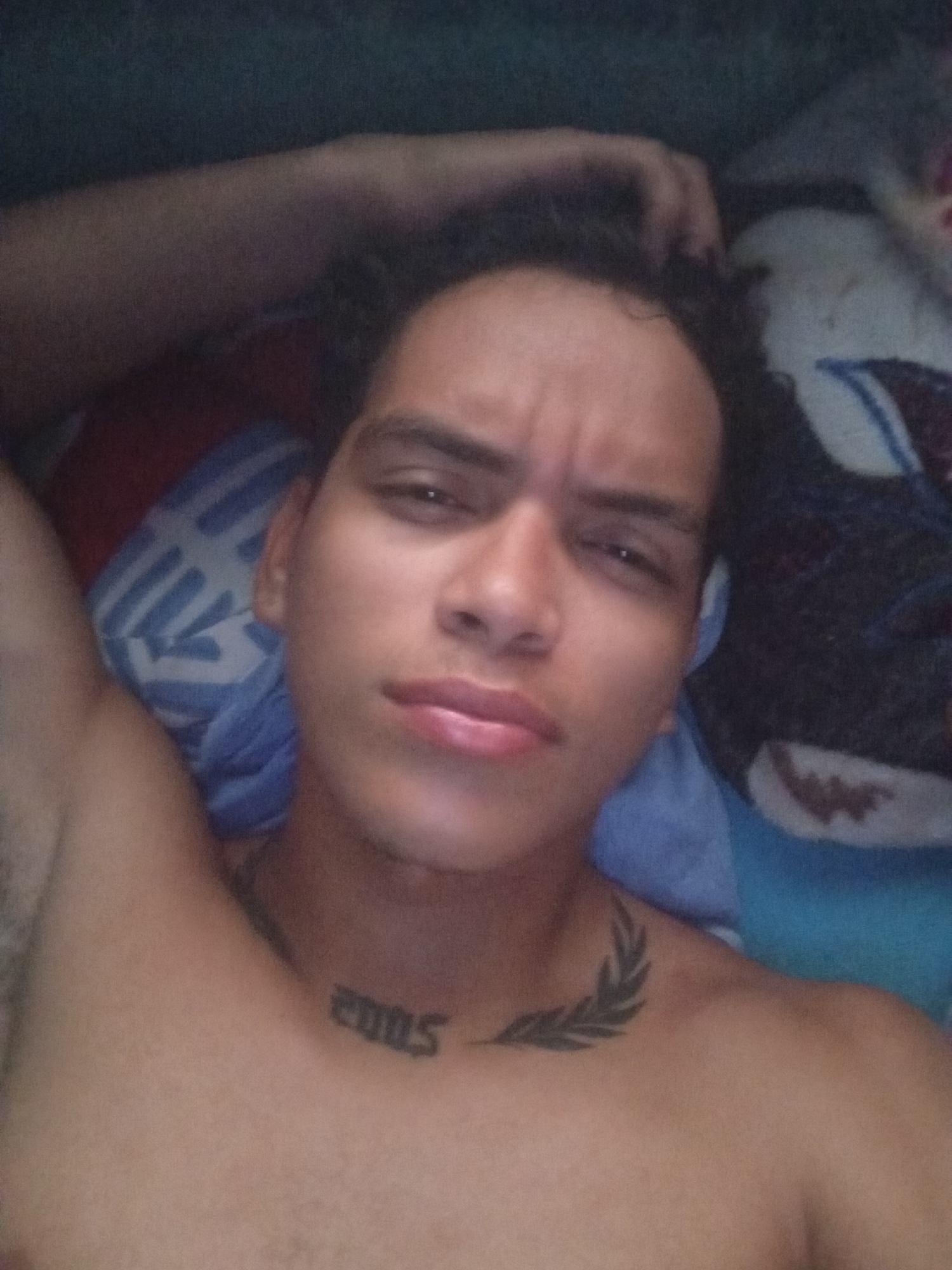 A selfie of a shirtless young adult male with brown curly hair. He has a slight frown. He has tattoos on his neck; the number 2005 and a laurel branch. He is lying in bed with patterned blue and white sheets. The location is unknown.