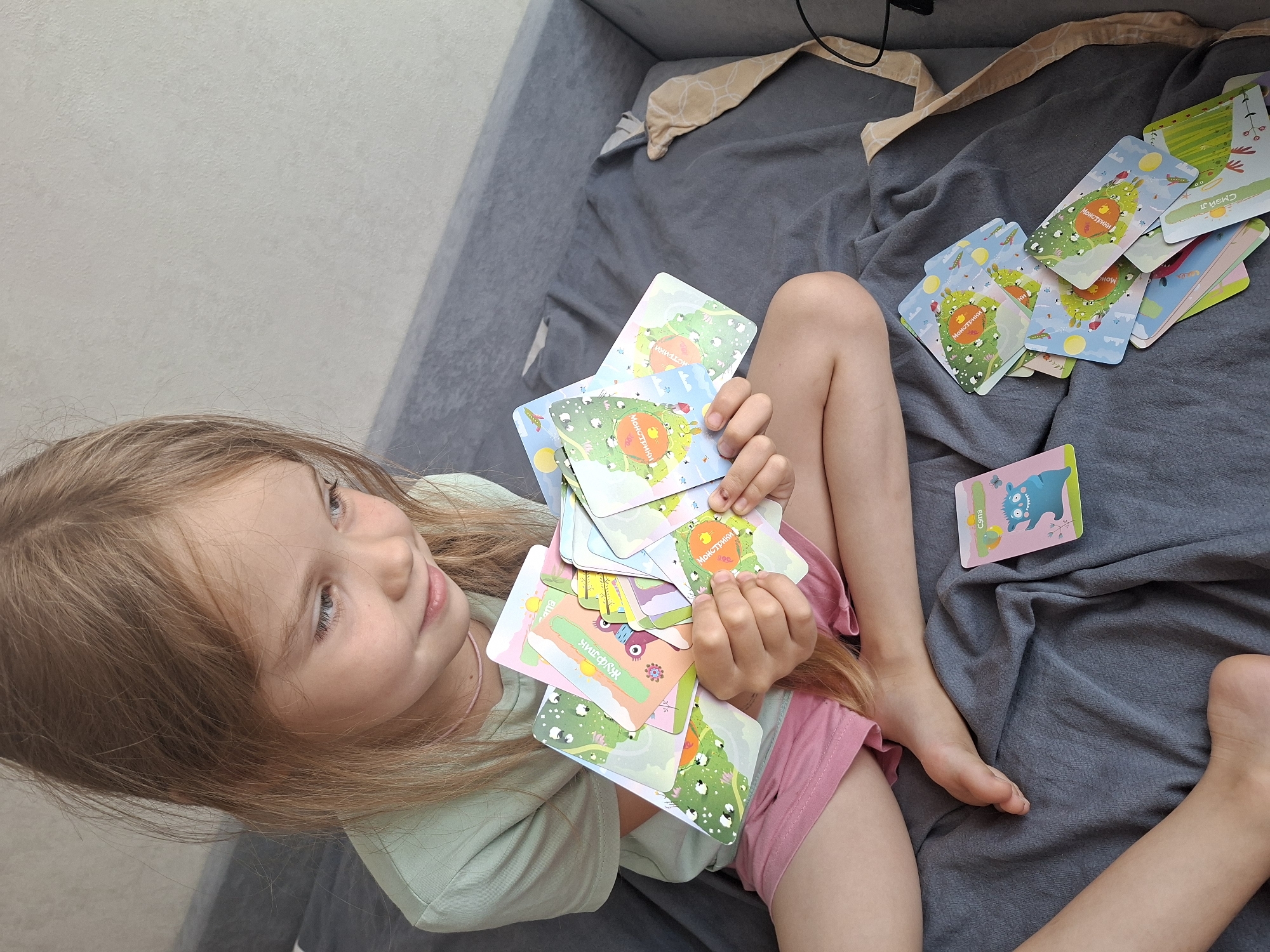 A young girl with long, blonde hair is sitting on a grey blanket, holding a pile of colorful cards. She is wearing a light green t-shirt and pink shorts. The cards feature cartoonish illustrations of monsters, sheep, and other whimsical elements, some with Cyrillic text. One card clearly visible shows a blue monster with the word "Сыма" written on it. Another card depicts a green monster with the word "МОНСТРИКИ" (Monsters) in Cyrillic.
The setting appears to be indoors, possibly on a bed or couch, given the soft grey fabric of the blanket and a hint of a grey upholstered surface in the background. The lighting suggests daytime. The girl's expression is one of engagement and curiosity as she looks at the cards. The scene is casual and suggests a moment of play or learning. The location context of Izhevsk, Russia, is noted.