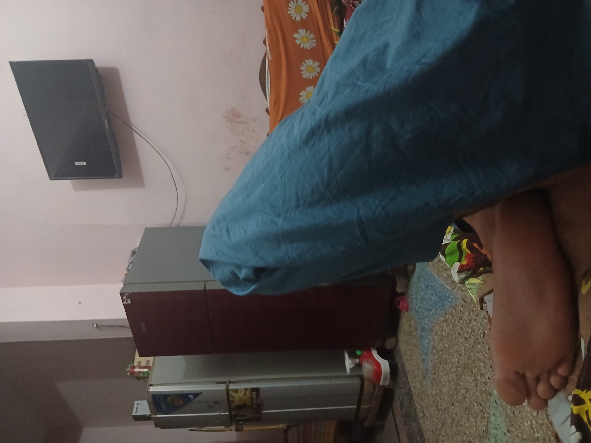 This is an indoor, eye-level, medium shot photograph taken in what appears to be a bedroom. The scene is captured in a low-light environment, with a dominant presence of a blue sheet or blanket covering a significant portion of the frame from the bottom right upwards and to the left. A bare human foot, with visible toes and sole, is positioned in the lower left corner, partially covered by patterned fabric.
In the background, to the left, are two refrigerators, one silver and one dark red, standing side-by-side. A small red and white toy car is placed on the floor in front of the refrigerators. Further to the left, a portion of a lighter-colored appliance, possibly another refrigerator or a cabinet, is visible. Above the refrigerators, a dark rectangular object, likely a television, is mounted on a light pink wall. The wall has some red marks near the top right. The floor appears to be tiled with a speckled pattern of brown and blue. An orange blanket with white daisy patterns is spread on a surface behind the blue sheet. The overall impression is a domestic setting within a residential space in Peshawar City Tehsil, Pakistan.