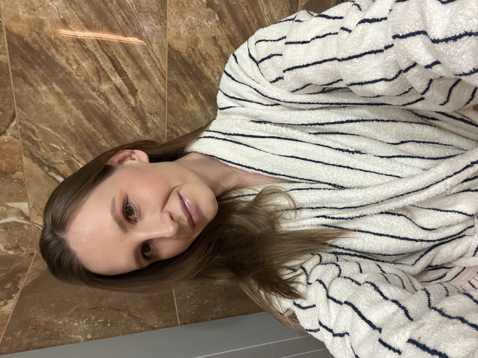 A female subject, appearing to be in her 20s or 30s with light brown hair and complexion, is depicted in a head-and-torso view, looking directly into the camera with a slight smile. She is dressed in a white terry cloth bathrobe featuring dark vertical stripes. The background consists of large rectangular brown marble-patterned tiles on the right, displaying a vertical light reflection, and a plain light grey wall section on the left. The scene takes place in an indoor environment, consistent with a bathroom or spa area. No specific visual identifiers are present to confirm the exact location within the city of Khimki, Russia.
