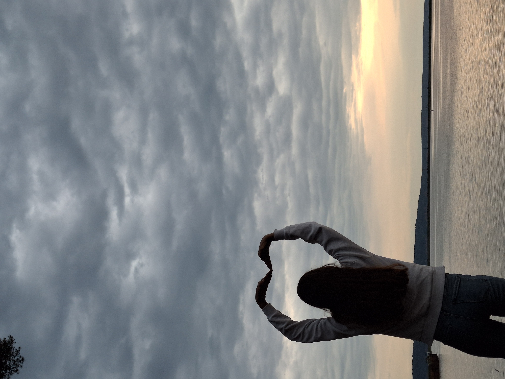 A young woman stands at the edge of a body of water, possibly the Kama River near Chaikovsky, Russia, indicated by the landscape and horizon in the background. She faces the water with her back to the viewer, making a heart shape with her hands above her head against a cloudy sky. She is wearing a light-colored hoodie and dark jeans. The water appears calm, with a light reflection on the surface. The sky is overcast, transitioning to a lighter hue near the horizon. A small portion of a tree is visible in the lower left corner of the frame.