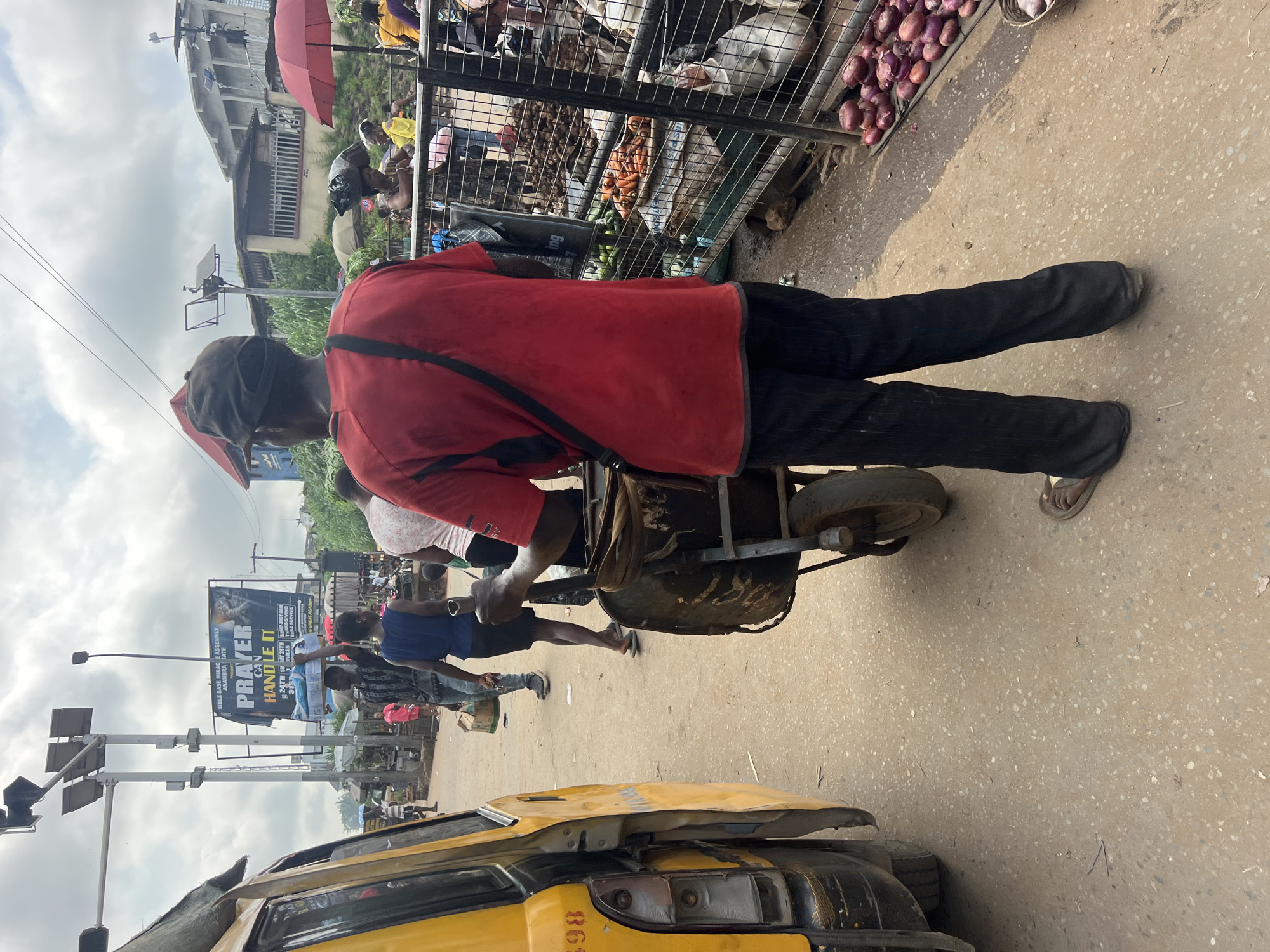 A busy street market scene unfolds in Nkpor, Nigeria, under an overcast sky. A man wearing a red t-shirt and black pants, with a black cap, pushes a wheelbarrow filled with items. He has a black strap across his chest. To his left, a yellow vehicle, possibly a taxi, is parked. In the background, various market stalls are visible, with vendors and customers interacting. Produce such as carrots and onions are displayed for sale, protected by metal fencing. Other individuals can be seen walking and engaging in market activities. A large billboard with text and images is also prominent in the background, suggesting a public announcement or advertisement. The ground is paved with asphalt, typical of a street in a Nigerian city.