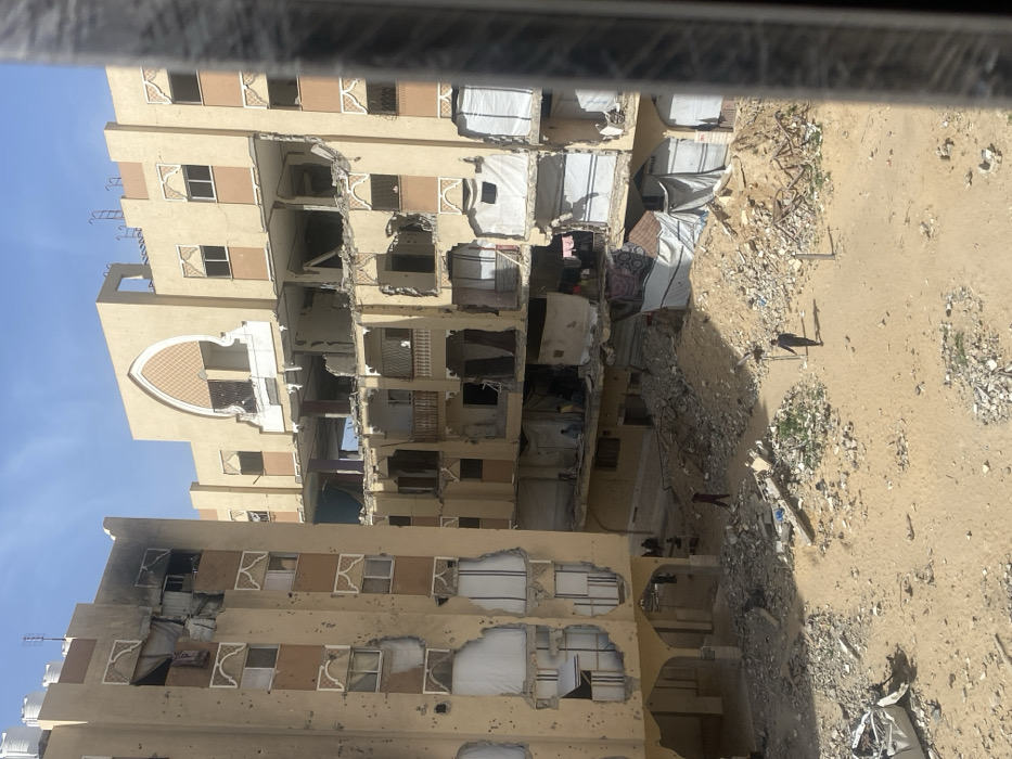 This image depicts a scene of extensive structural destruction in Khan Yunis, Palestinian Territory, under clear daylight conditions. Two multi-story residential buildings, primarily beige in color, exhibit severe damage. The building on the left features numerous impact holes across its facade, destroyed windows (some covered with white material), and significantly damaged upper sections with exposed rebar. Burn marks are visible on the facade near the top-left, and white water tanks are present on its roof. The building on the right, partially visible behind and to its right, shows catastrophic structural failure, with multiple floors partially collapsed or missing, exposing interior spaces. Some original decorative arched window frames remain on its upper sections. White tarps or makeshift coverings are draped over various openings on both structures, particularly evident within the damaged lower levels of the right building, suggesting temporary enclosures.
The ground area between and in front of the buildings is an open expanse of sand and dirt heavily covered with concrete rubble, twisted metal, and general building debris, with scattered piles of refuse. Approximately seven individuals are visible within the scene. Two men walk across the rubble-strewn ground in the foreground; one in a dark shirt carries a stick or rod, and another in a dark red shirt walks towards the left building. At least five other individuals are discernible within the lower, exposed levels of the damaged right building, some appearing to be seated or standing amidst the debris. No direct interactions between individuals in the foreground are clearly visible.