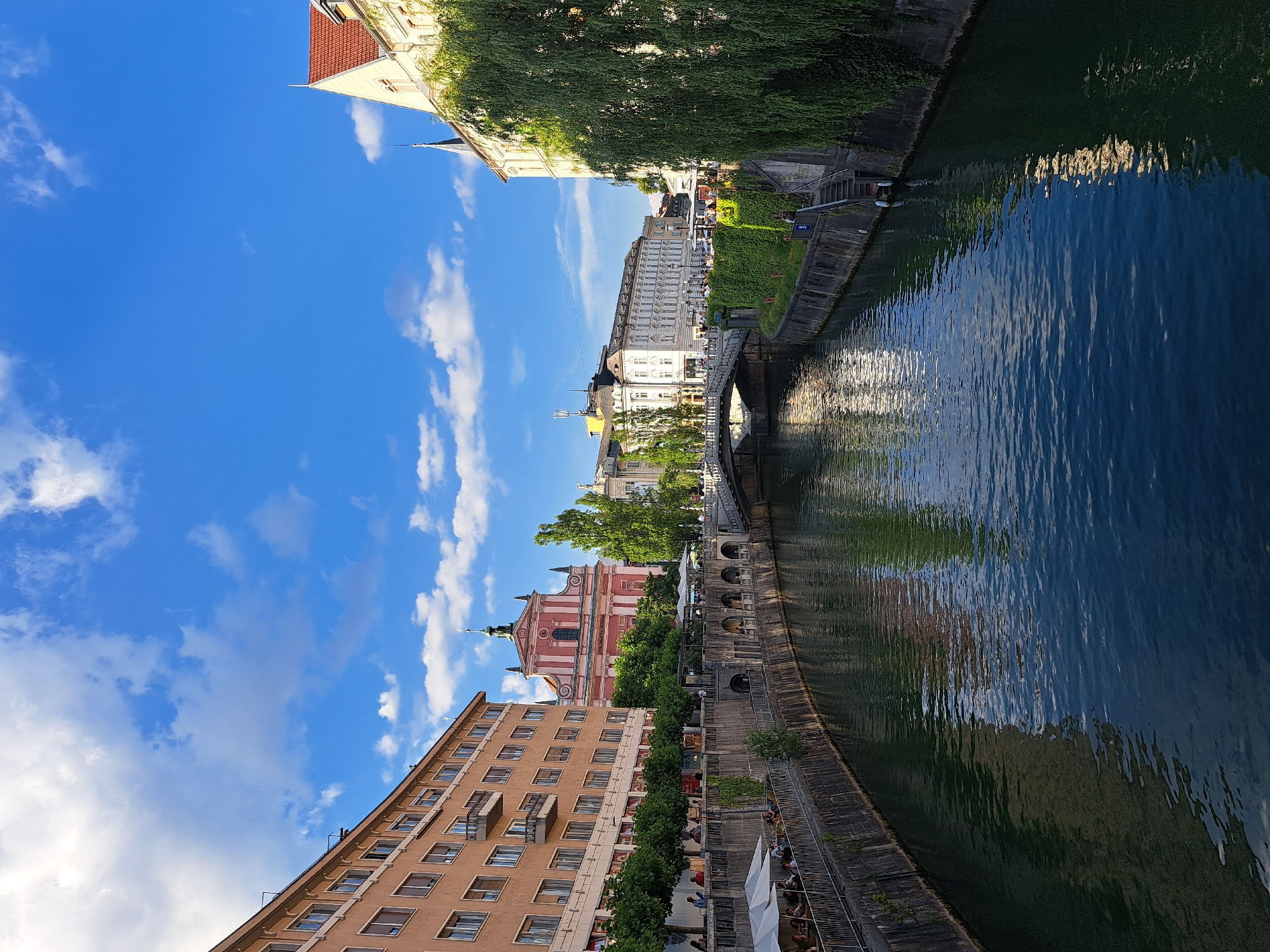 The image shows a view of a canal in a European city, likely Ljubljana, Slovenia, on a sunny day with a blue sky and scattered clouds. The canal is lined with buildings on both sides. On the left, there is a multi-story building with a salmon-colored facade and many windows. A walkway with outdoor seating and umbrellas runs along the canal in front of this building.
Across the canal, a pink church with a green dome and spires is prominently visible. Further along the canal, more buildings with white facades and intricate architectural details can be seen. A stone bridge arches over the canal, with people walking across it. Lush green trees and ivy cover parts of the canal walls and buildings, adding to the picturesque scene. The water in the canal is a deep blue, reflecting the sky and the surrounding architecture. Several people are visible on the walkways and the bridge, enjoying the day.