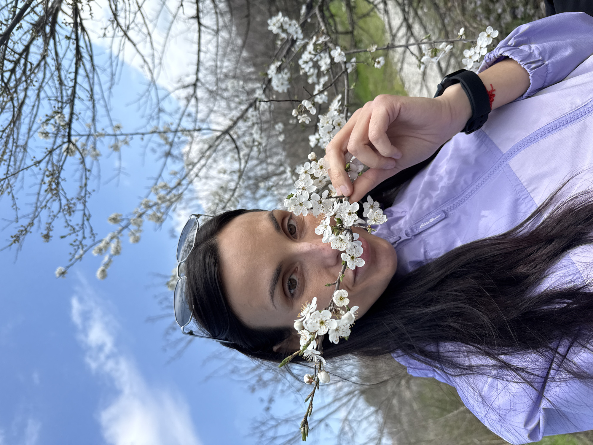 The image shows a woman holding a flowering branch in front of her face, seemingly outdoors. She is wearing a light purple jacket and has sunglasses perched on her head. A black sports watch is visible on her wrist, with a small red string or bracelet nearby. The background includes other flowering trees, a blue sky with some clouds, and hints of a green landscape. There is no way to know from this image whether or not the location is in the city of Pleven, Bulgaria.