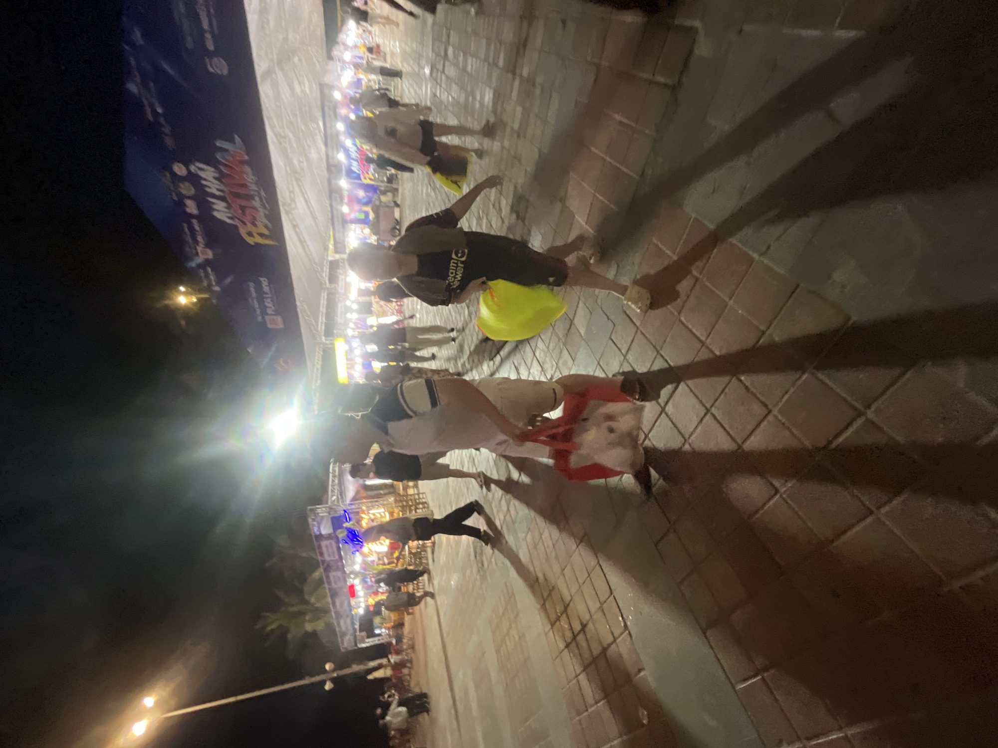 The image depicts an outdoor night scene, likely a street or promenade, in Da Nang, Vietnam, during a festival, as indicated by the banner that reads "AN HAI FESTIVAL". The foreground is dominated by a tiled walkway, reflecting the ambient light. Several individuals are visible, most of whom are walking.
In the mid-ground, a man in a white shirt with blue accents and beige shorts is walking towards the left, carrying a red tote bag that appears to contain white items, possibly stuffed animals. To his right, a person wearing a dark t-shirt with "Teamviewer" text and dark shorts is walking towards the viewer, holding a bright yellow bag in their left hand.
In the background, stalls and tents are set up, illuminated by bright lights, suggesting a marketplace or event area. Palm trees are faintly visible behind the structures. A lamppost with multiple lights stands on the left side of the image. The overall atmosphere is lively, characteristic of a nighttime festival.