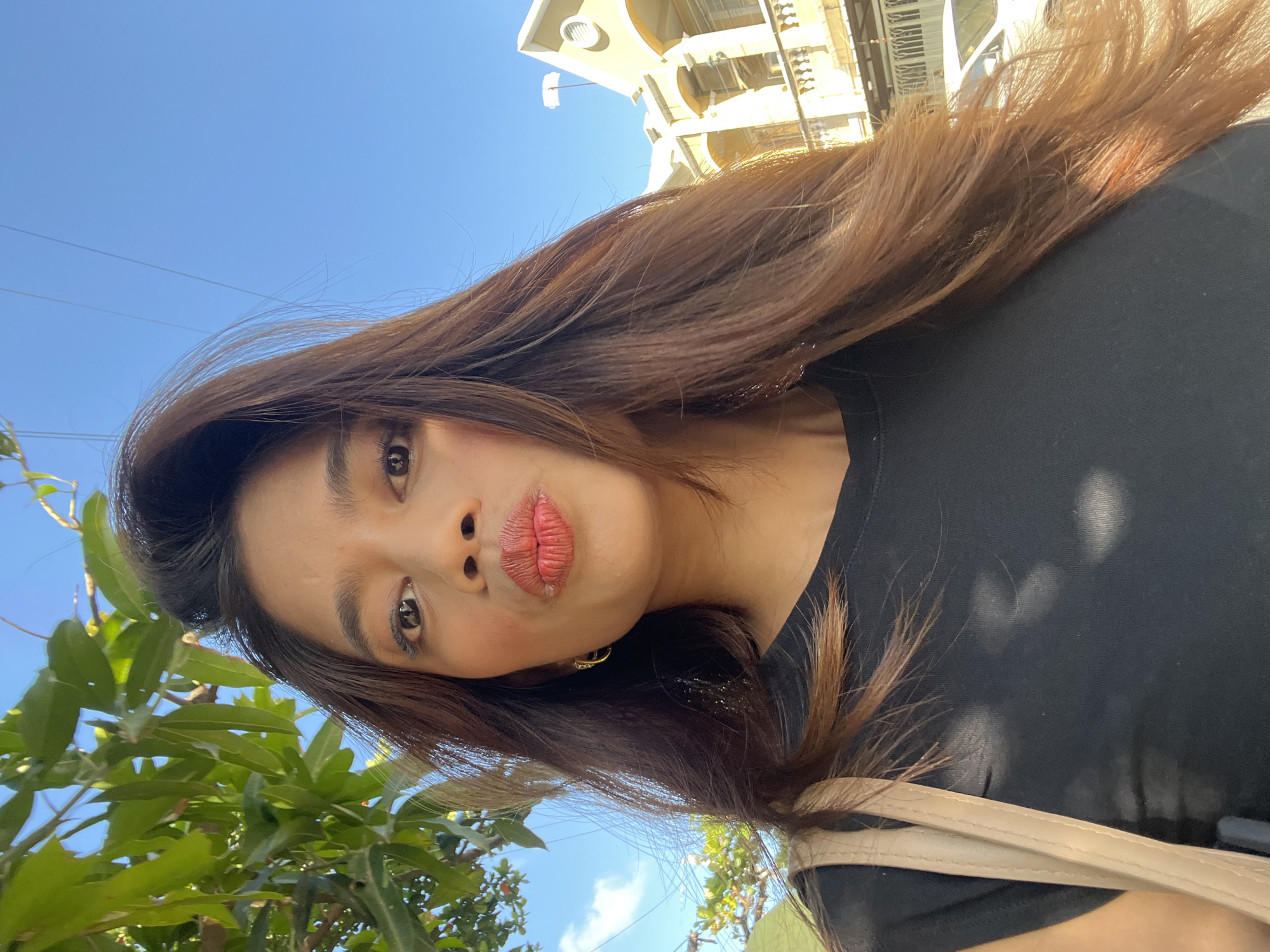 In this selfie, a young woman with long, dark brown hair and bright red lipstick pouts her lips. She is wearing a black t-shirt and a light-colored shoulder bag. Sunlight casts dappled shadows on her shirt. In the background, lush green leaves of a tree are visible to the left, and a building with arched windows and a tiled roof, characteristic of architecture in Davao City, Philippines, stands to the right. The sky is a clear blue with a few wispy clouds. The scene suggests an outdoor setting during the day in a tropical urban environment.