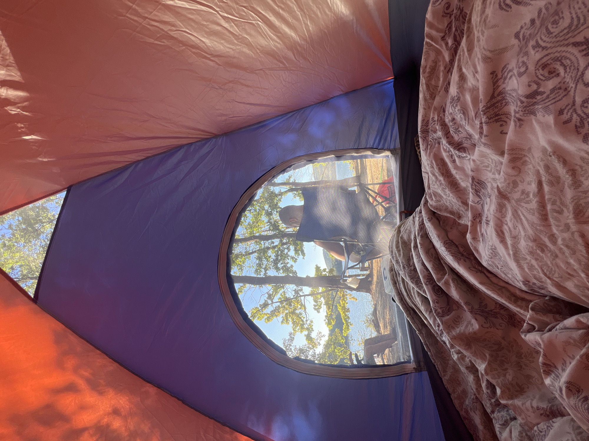 An interior view from inside a tent looking out through an arched opening at a lakeside scene. The tent interior is colorful with orange and purple fabric visible, and a patterned comforter fills the foreground. Outside the tent, a person sits in a camping chair facing away from the camera, towards a body of water with trees on the opposite bank. Sunlight filters through the leaves of the trees, casting shadows on the ground. The environment appears to be a campsite near a lake.