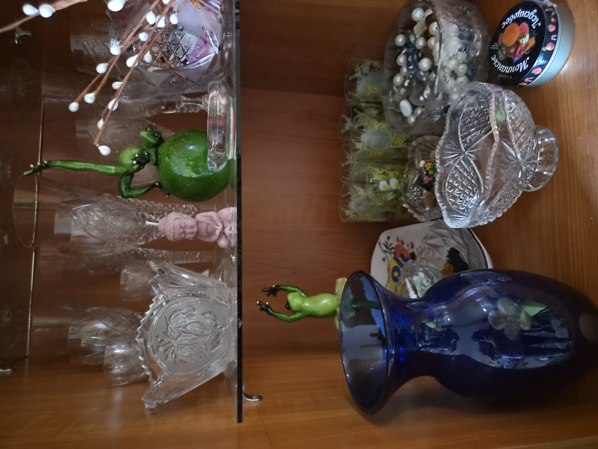 The image shows a wooden cabinet with glass shelves displaying various decorative objects. On the top shelf, there are several crystal glasses, a large crystal bowl with a floral pattern, a small pink Buddha figurine, a green frog figurine, and a decorative sprig with white bead-like elements. On the bottom shelf, there is a large dark blue glass vase, a patterned plate, several small decorative glasses, a crystal bowl filled with beads and pearls, another crystal bowl, and a round tin with fruit imagery. The overall environment appears to be an indoor setting, possibly a display cabinet in a home or shop. There is no identifiable location in the city of Arseniev, Russia, or any specific details about people, their actions, or interactions.