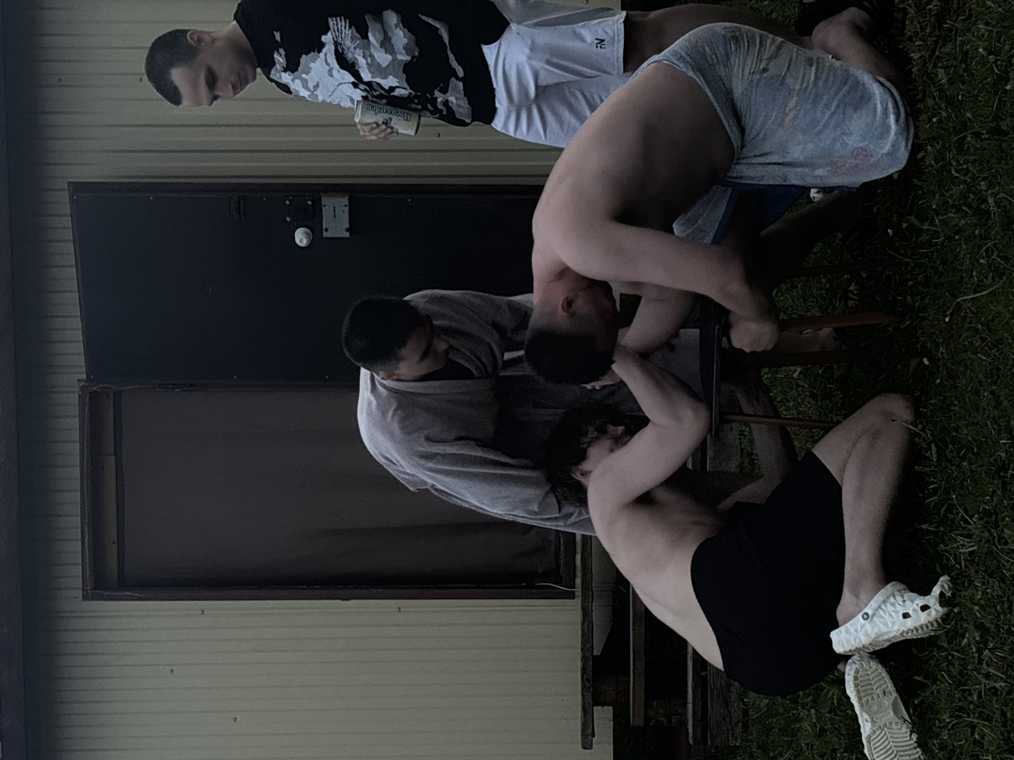 In Пальники, Russia, the image depicts four individuals outdoors near a light-colored building with a dark door and a window frame. One person is standing and holding a beverage can, wearing a black and white patterned top and white shorts. Another individual is seated, wearing patterned blue shorts. Two other people are positioned closer to the building, with one wearing a gray robe and the other wearing black shorts and white shoes. The individuals appear to be interacting around a chair. The scene is situated on grass.
