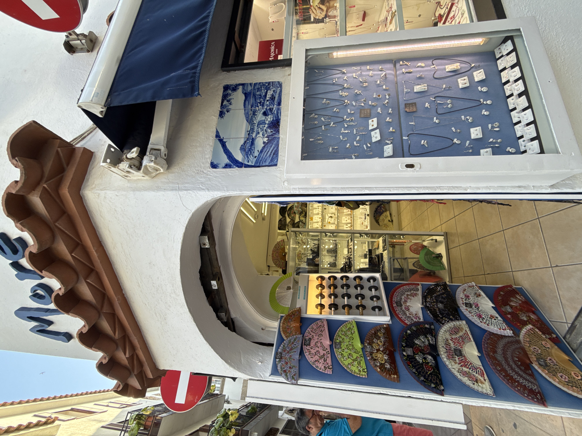 This media file depicts a street scene in Tossa de Mar, Spain, likely during daylight given the bright lighting and clear skies. The focus is on a shopfront displaying various items for sale.
The primary subjects are the merchandise and the shop's exterior. In the foreground, a table is laid out with an assortment of decorative fans, many featuring intricate floral patterns in vibrant colors like pink, green, red, and brown, as well as darker, more subdued designs. Behind the fans, a display case showcases a collection of jewelry, including necklaces, earrings, and pendants, arranged on a blue background. The jewelry appears delicate and possibly made of silver.
The shop itself has a white facade, with a blue awning extending over a section of the entrance. A decorative tiled panel, depicting a blue and white landscape scene, is mounted on the wall. The entrance to the shop leads into a space where more merchandise is visible, including what appears to be additional jewelry displays.
Notable details include a brown, decorative architectural element on the building's exterior, possibly a cornice or a decorative molding. A red and white circular sign is partially visible on the left side of the image. The overall atmosphere suggests a sunny day in a Mediterranean tourist town, with an emphasis on artisanal crafts and souvenirs. There is a glimpse of people in the background, one individual wearing a blue t-shirt. The image is taken from a slightly elevated, angled perspective, looking downwards and across the scene.