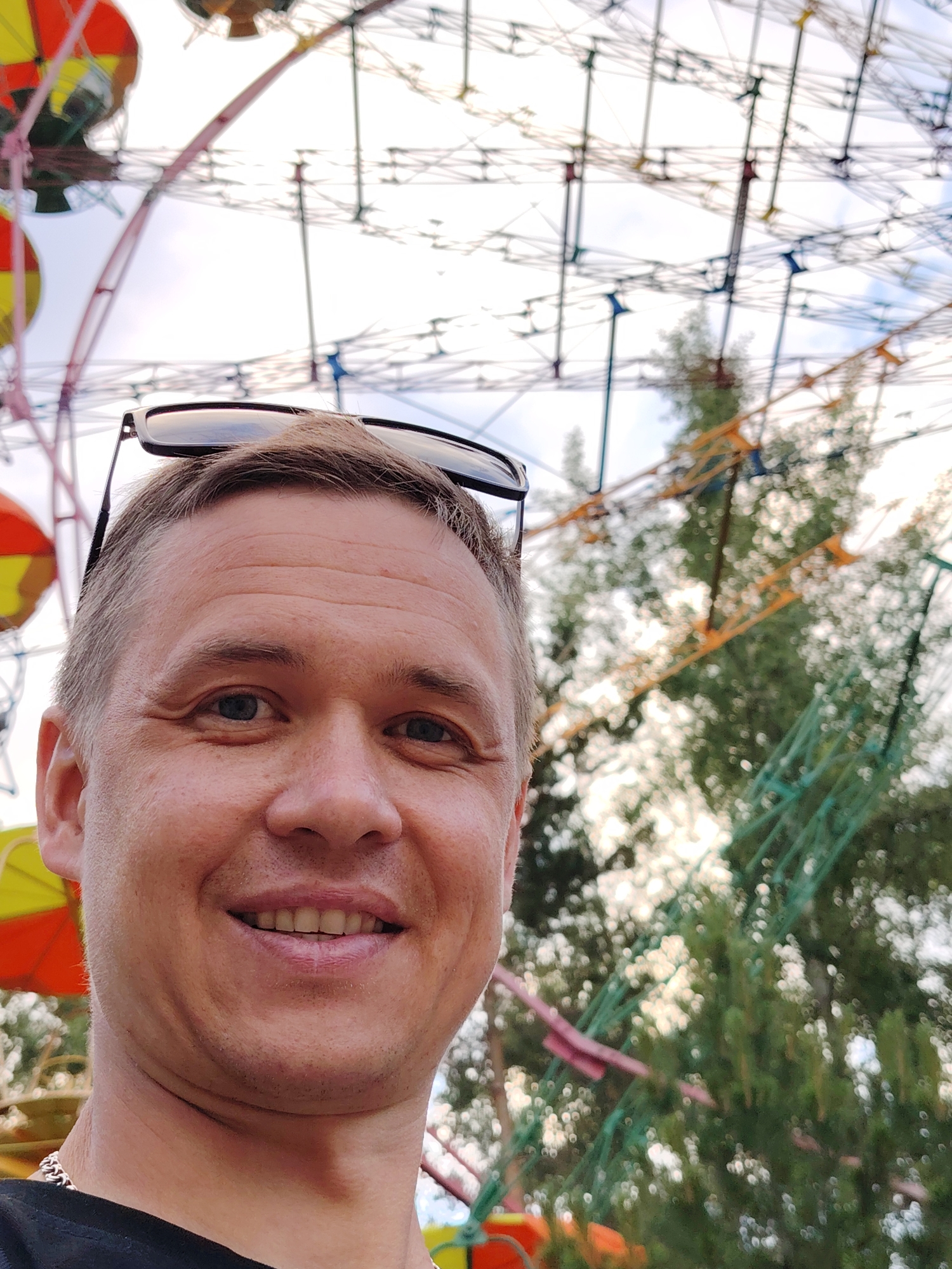 A man with short brown hair and sunglasses pushed up on his head takes a selfie outdoors. He is wearing a black t-shirt and a silver chain. Behind him, blurred elements of a Ferris wheel and trees are visible. The scene appears to be in a park or amusement area in Chaikovsky, Russia.