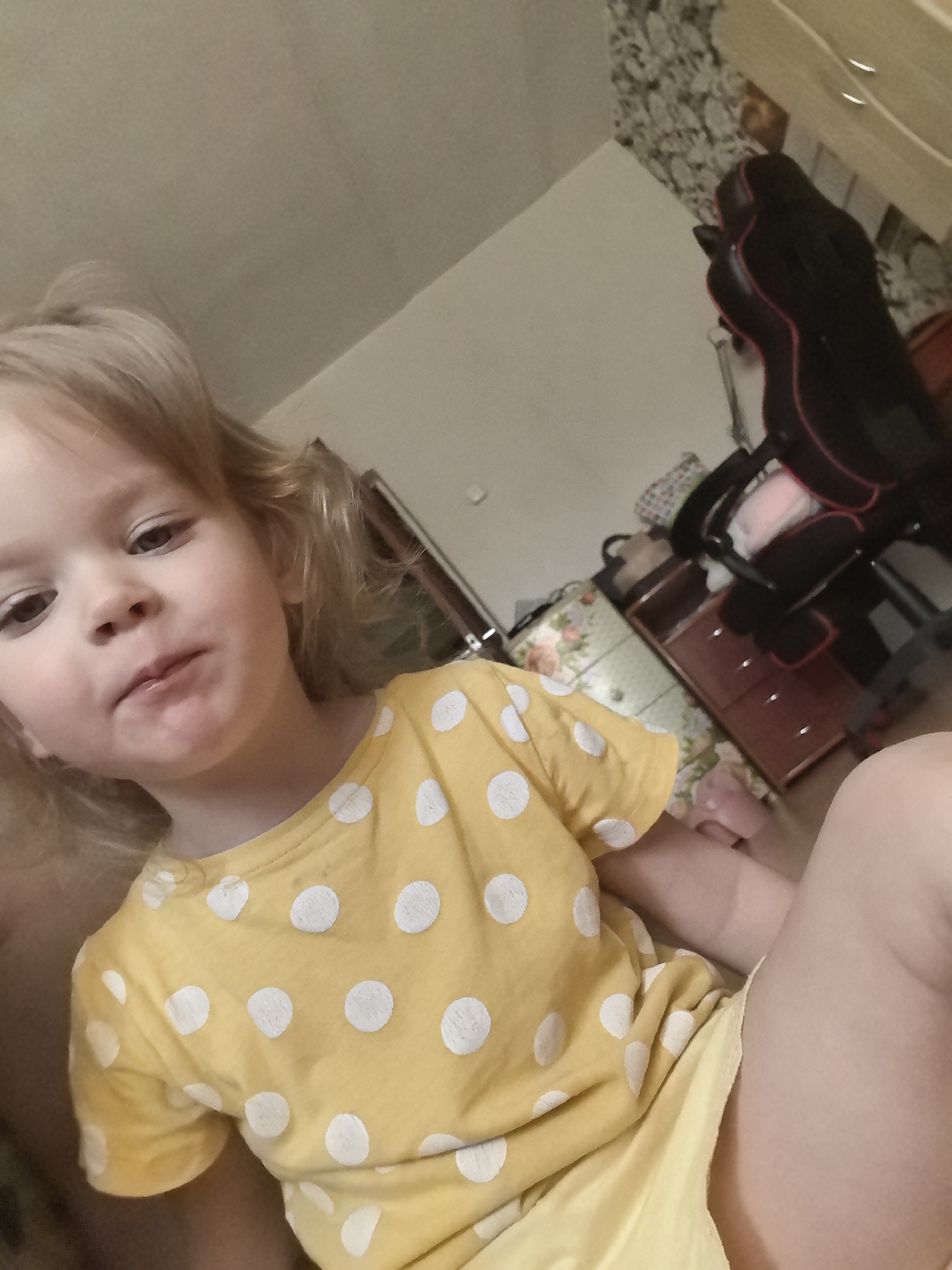 A child is lying down in a room and looking at the camera. The child is wearing a yellow t-shirt with white polka dots. The background of the image shows a dresser, a chair, and a wall. The room appears to be a bedroom. The location is unknown.